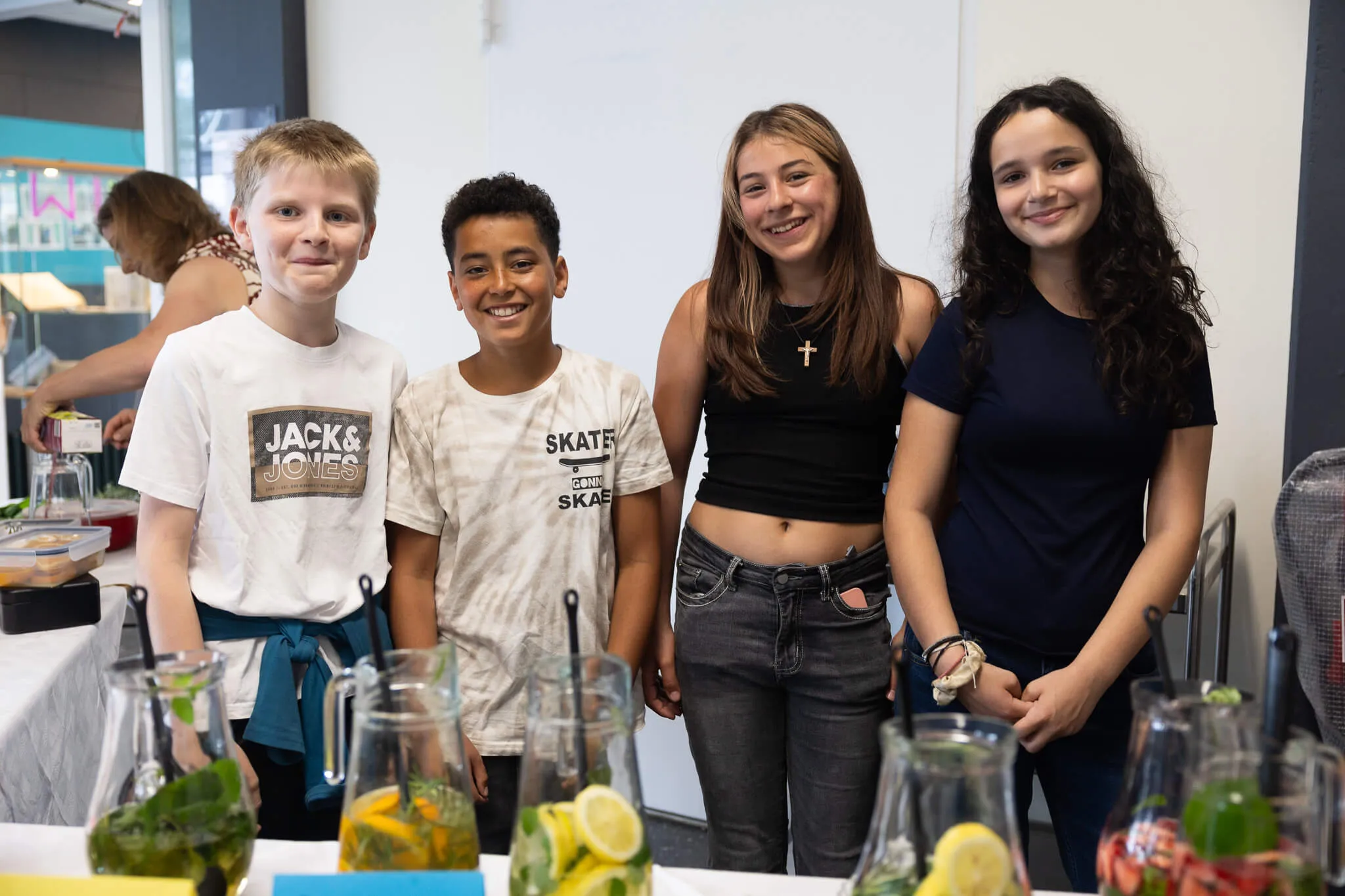 Vier lächelnde Kinder stehen hinter einem Tisch mit bunten Getränken. Im Hintergrund sind weitere Personen und ein Schulfest zu sehen.