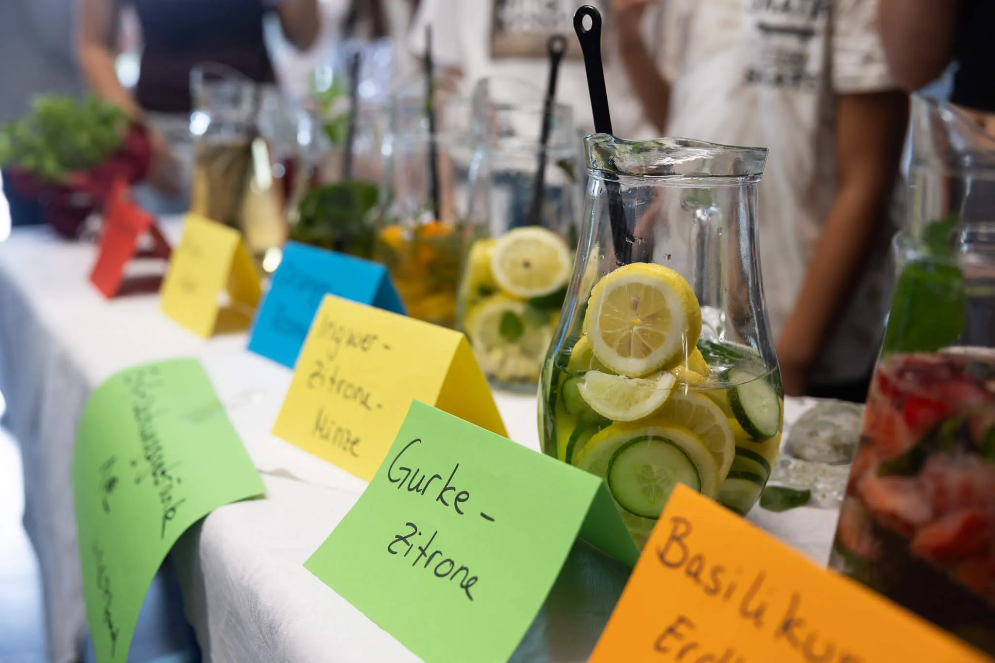 Tisch mit verschiedenen Getränkespendern und Beschriftungen wie „Gurke-Zitrone“ und „Ingwer-Zitronen-Minze“ auf einem Schulfest.