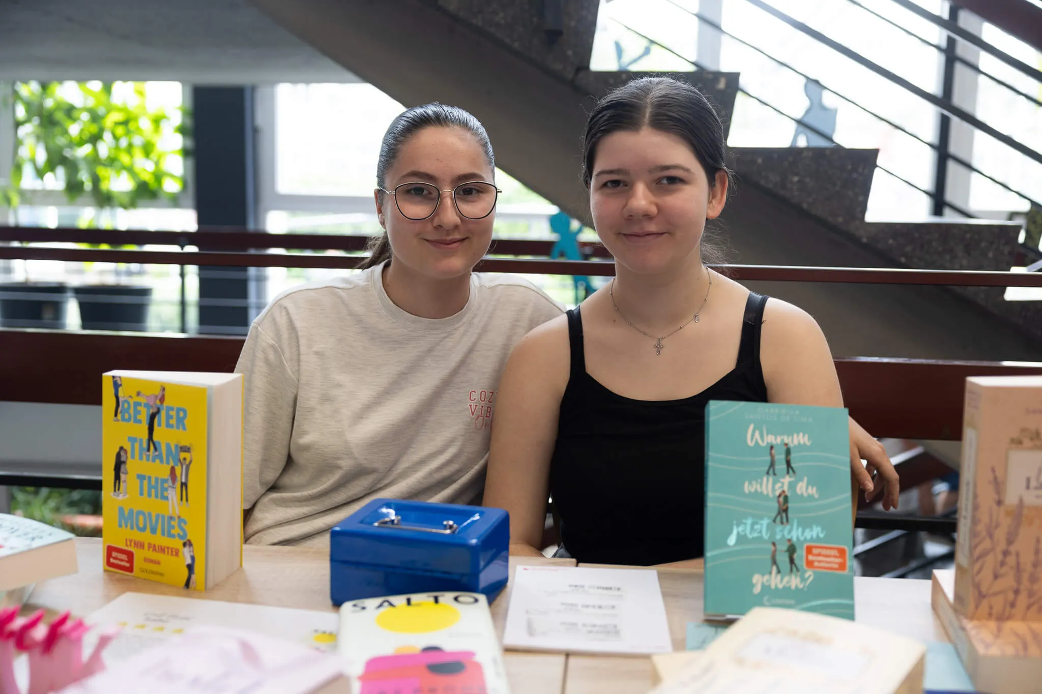 Zwei junge Frauen sitzen an einem Tisch mit Büchern und einer Spardose, lächeln in die Kamera. Im Hintergrund sind weitere Personen und ein festlich dekorierter Raum zu sehen.