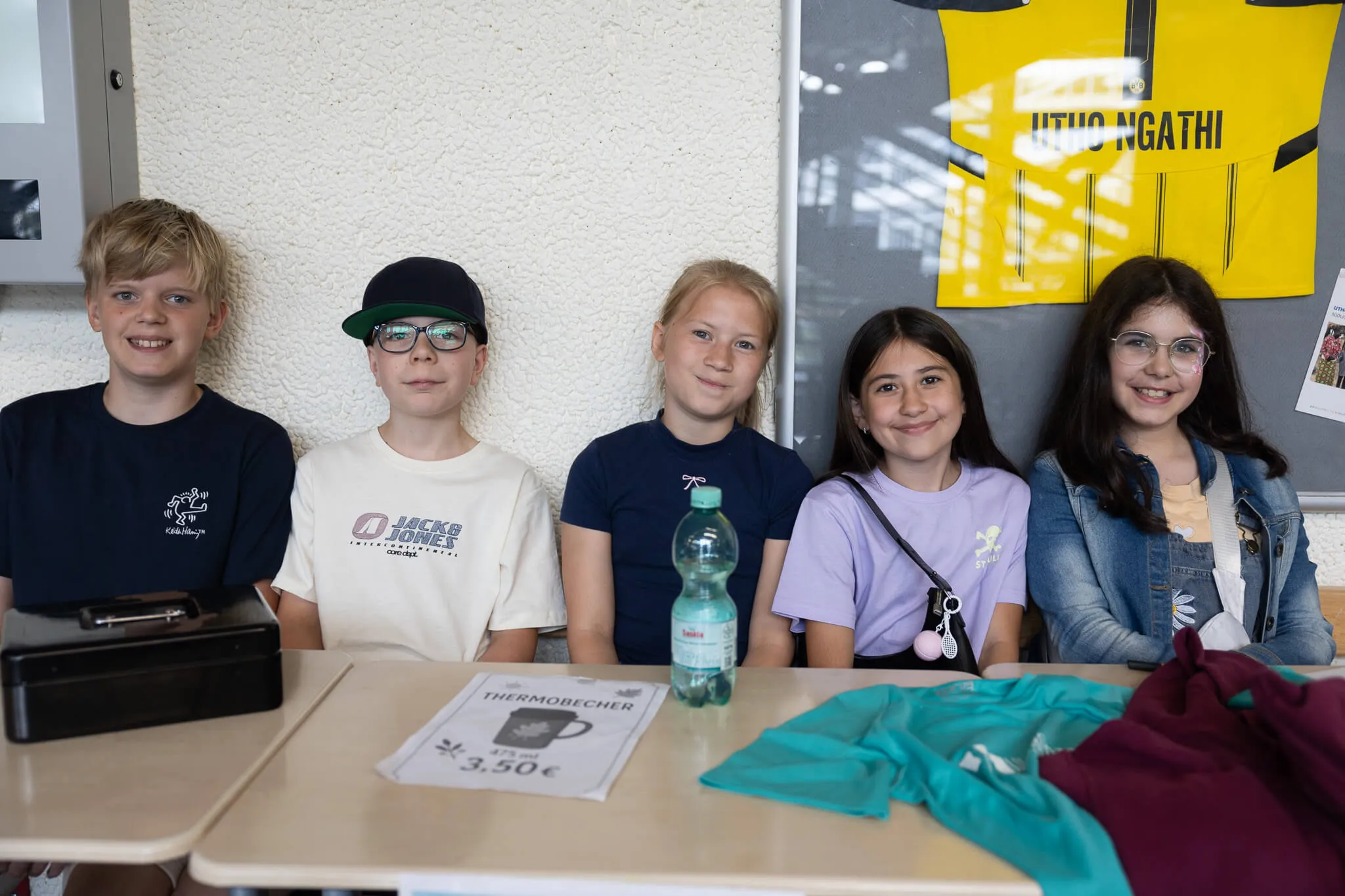 Fünf Schüler stehen nebeneinander an einem Tisch, lächeln in die Kamera. Im Hintergrund ist ein Plakat mit der Aufschrift „Hito Ngathi“ zu sehen.