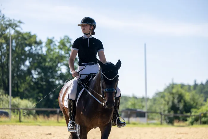 Eine Reiterin in schwarzer Kleidung und Helm sitzt auf einem dunklen Pferd und reitet auf einem sandfarbenen Platz. Im Hintergrund sind Bäume und ein blauer Himmel zu sehen.