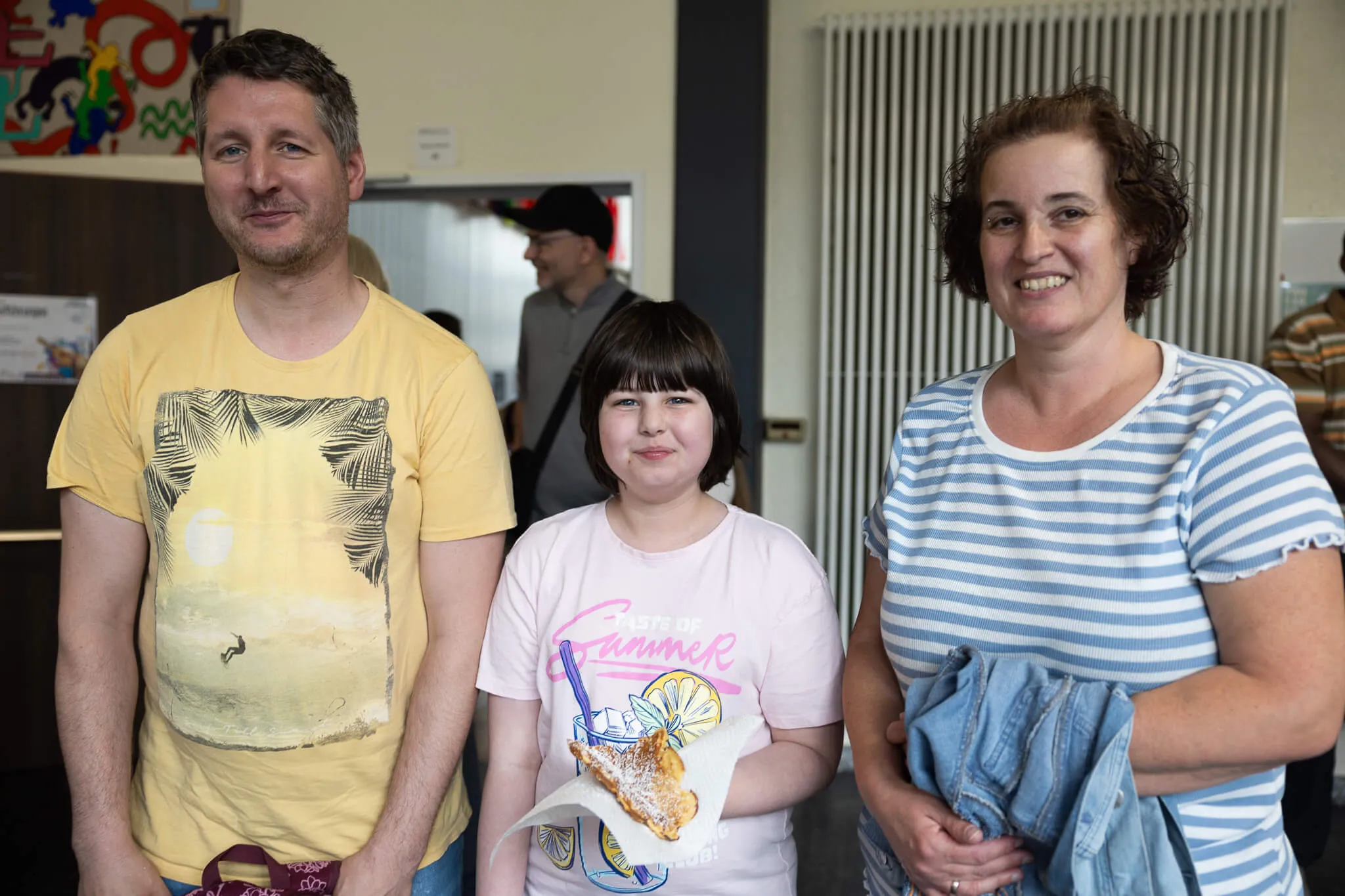 Vater, Tochter und Mutter stehen lächelnd vor der Kamera. Die Tochter hält einen frisch gebackenen Pfannkuchen in der Hand.
