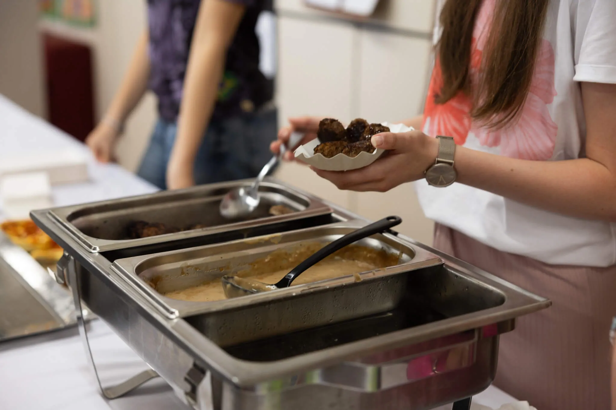 Ein Buffet mit Speisen steht auf einem Tisch, eine Person reicht einen Teller mit kleinen Fleischbällchen weiter.