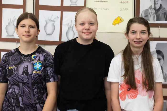 Drei junge Frauen stehen nebeneinander vor einer Wand mit Bildern und Preisschildern. Links trägt eine Brasilien-Trikot, die anderen tragen Freizeitkleidung.