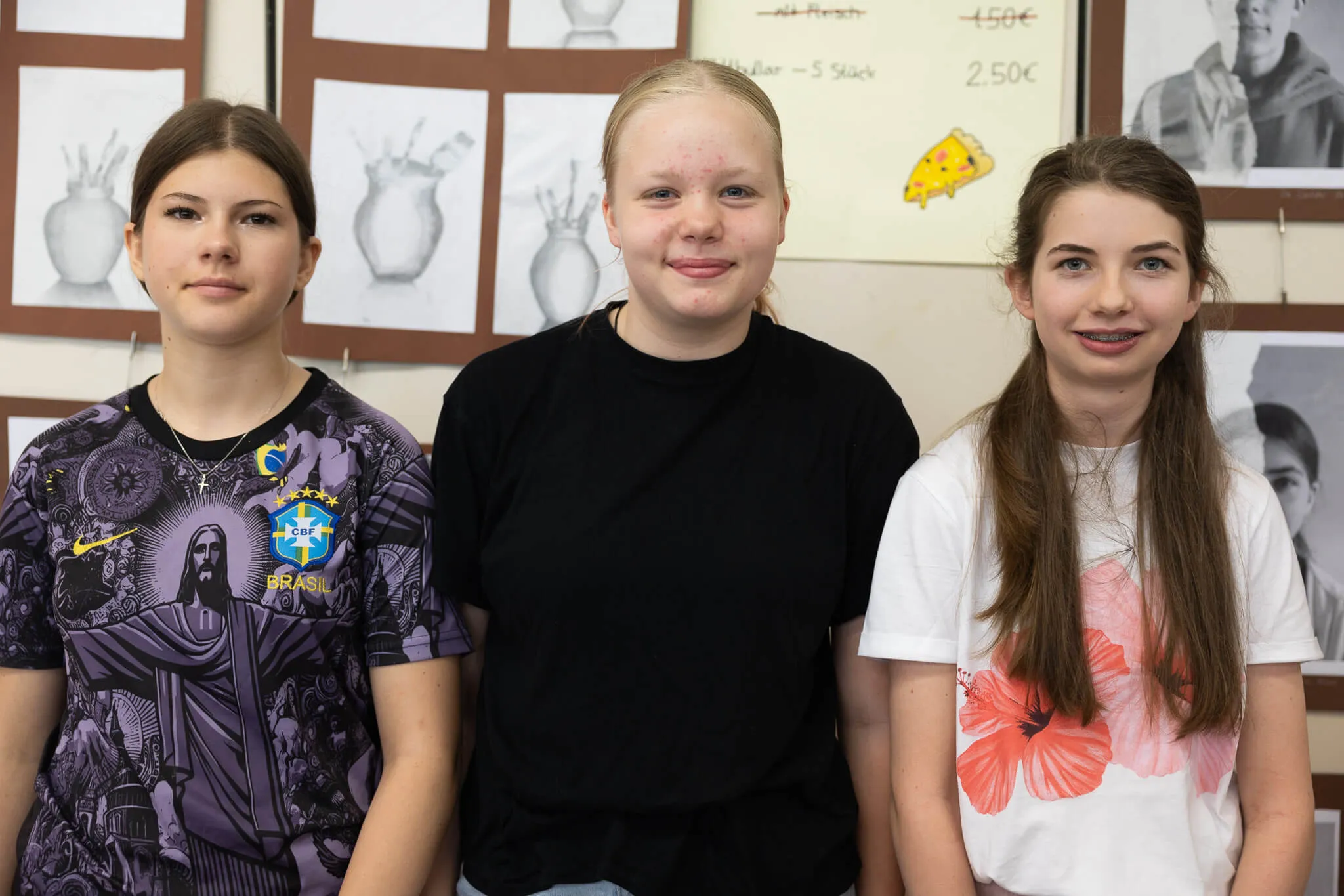 Drei junge Frauen stehen nebeneinander vor einer Wand mit Bildern und Preisschildern. Links trägt eine Brasilien-Trikot, die anderen tragen Freizeitkleidung.