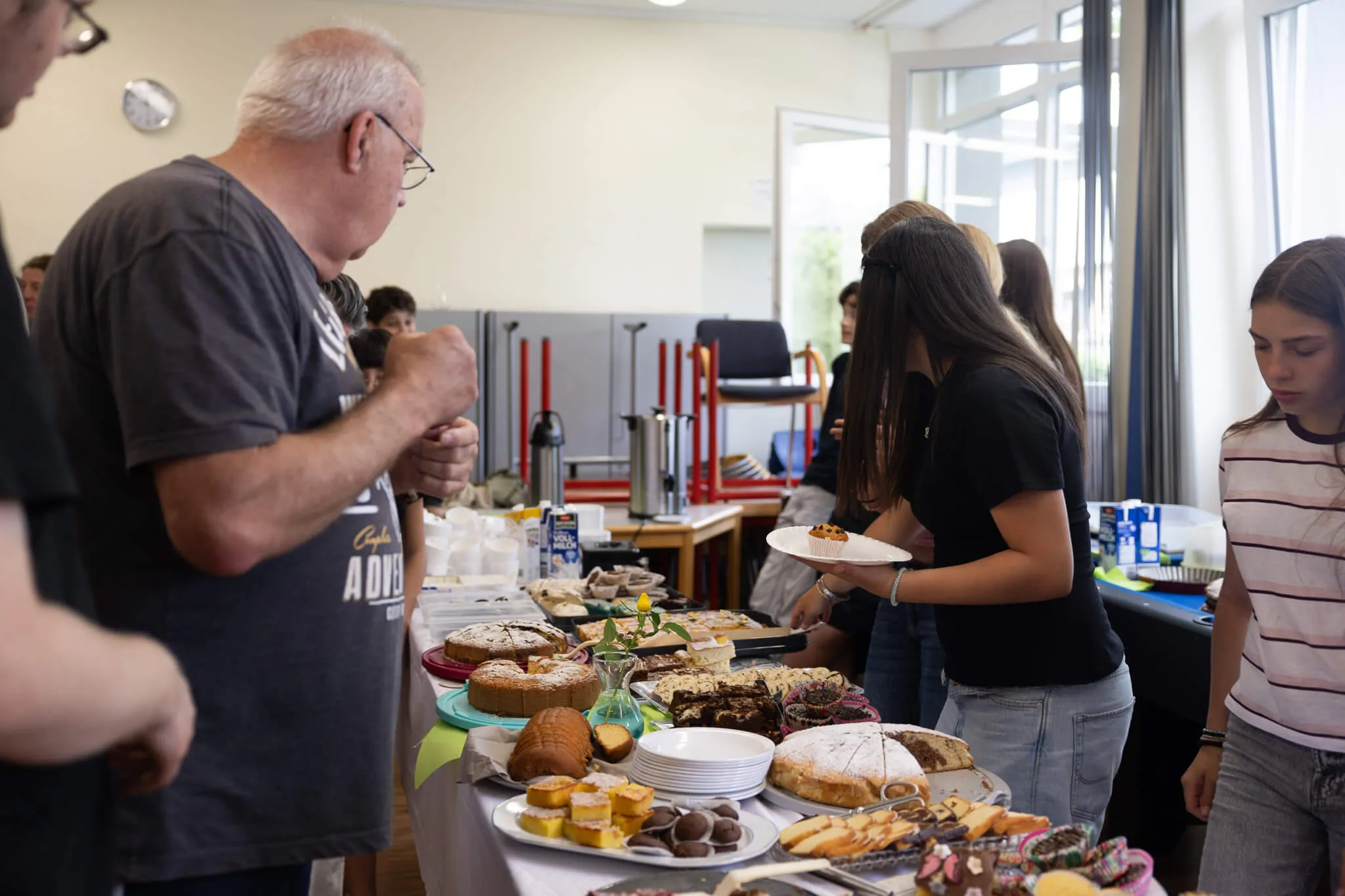 Ein Mann nimmt Kuchen von einem Tisch mit einer Auswahl an selbstgebackenen Torten und Gebäck, während eine junge Frau ihm ein Stück anbietet. Im Hintergrund stehen weitere Personen auf einem Schulfest.