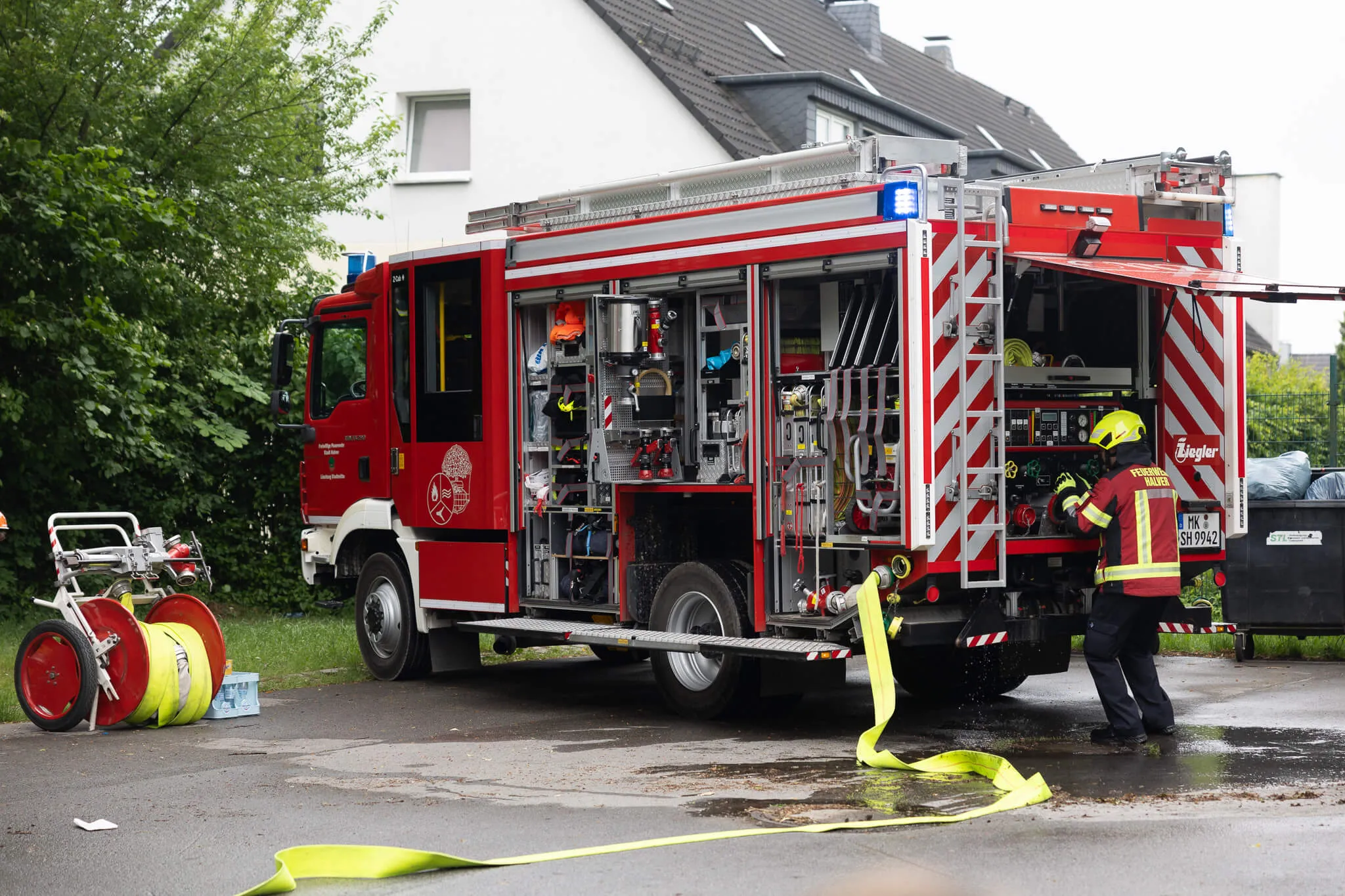 Roter Feuerwehrauto mit geöffneten Fächern und ausgefahrenem Schlauch vor einem Wohnhaus.