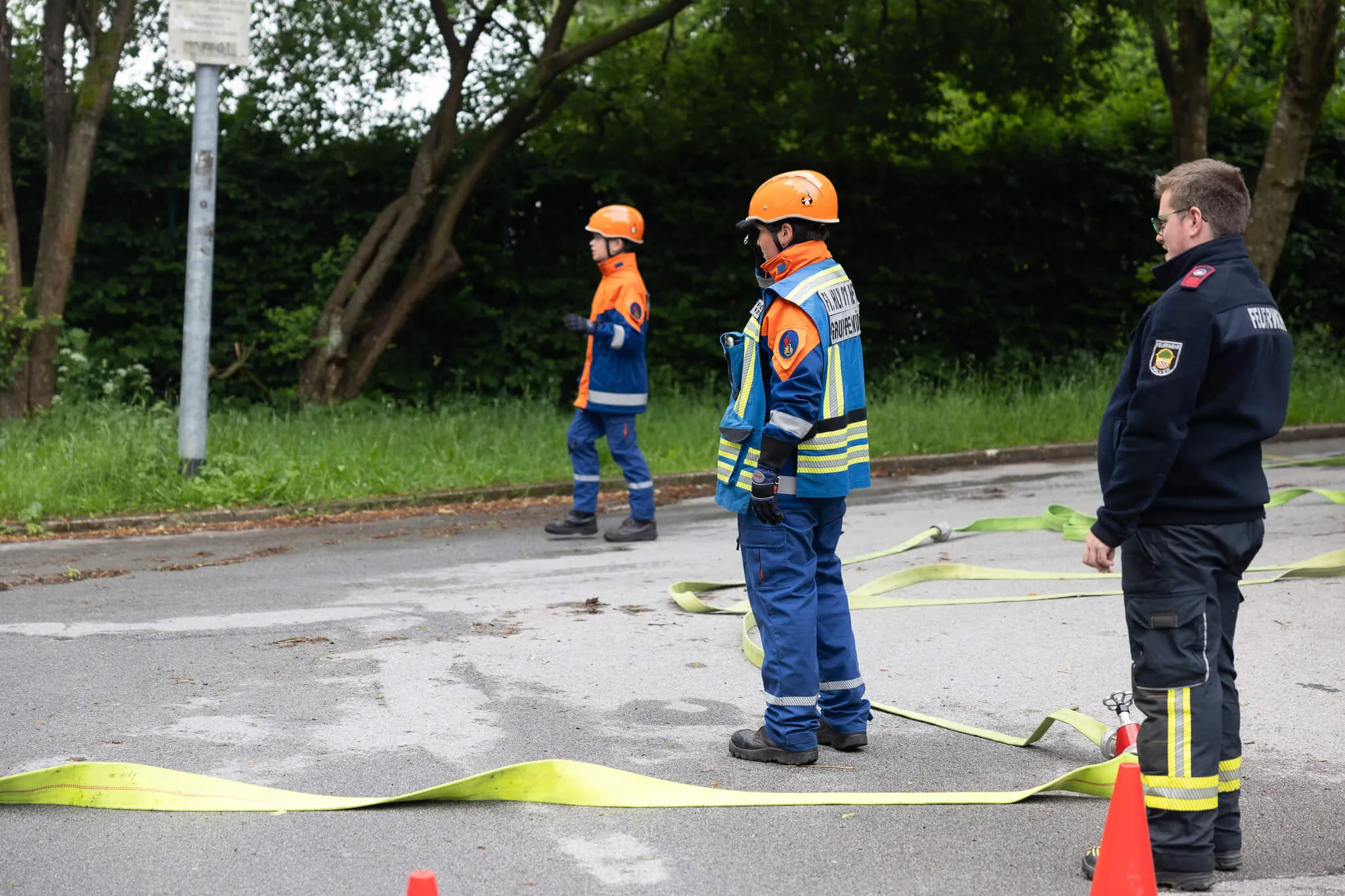 Vier Feuerwehrleute in Einsatzkleidung und Helmen stehen auf einem Parkplatz neben einem gelben Schlauch. Im Hintergrund sind Bäume und ein Verkehrsschild zu sehen.