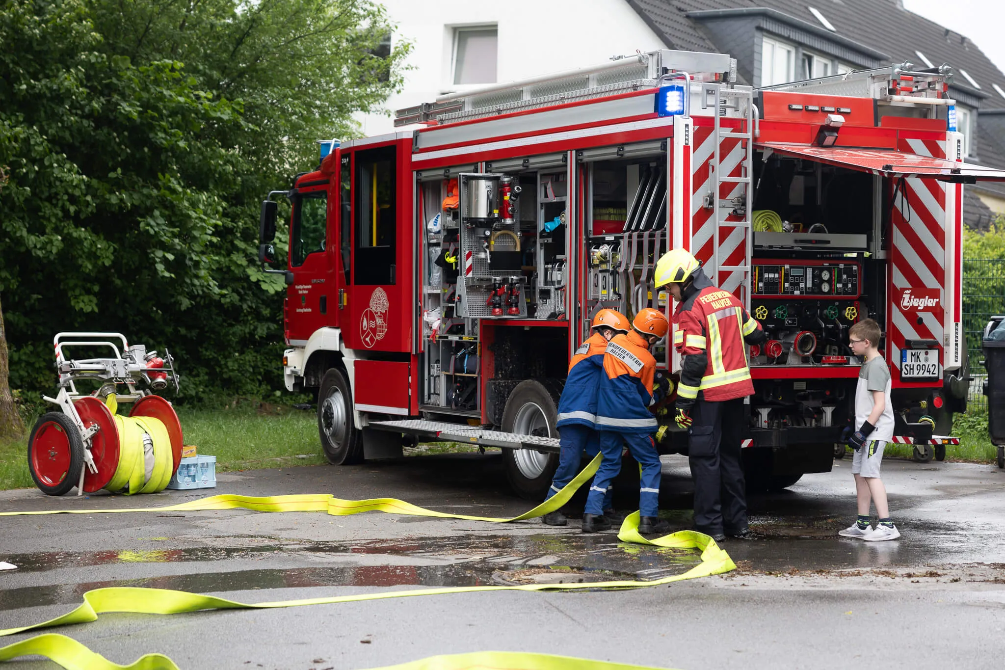 Feuerwehrleute bei der Arbeit vor einem roten Feuerwehrauto, während Kinder zusehen. Im Hintergrund ein Wohnhaus und Bäume.