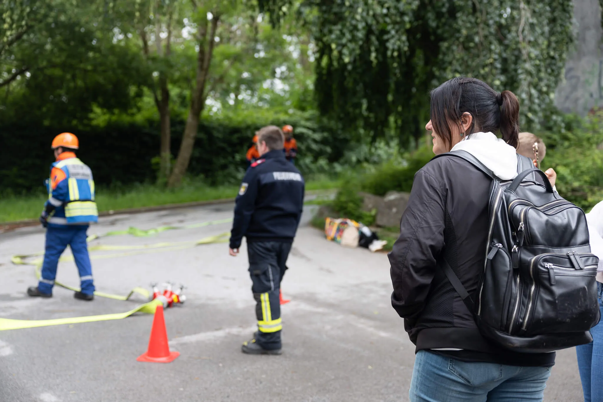 Eine Frau mit Rucksack steht vor Einsatzkräften und Absperrband an einer Straße, im Hintergrund Bäume und Büsche.