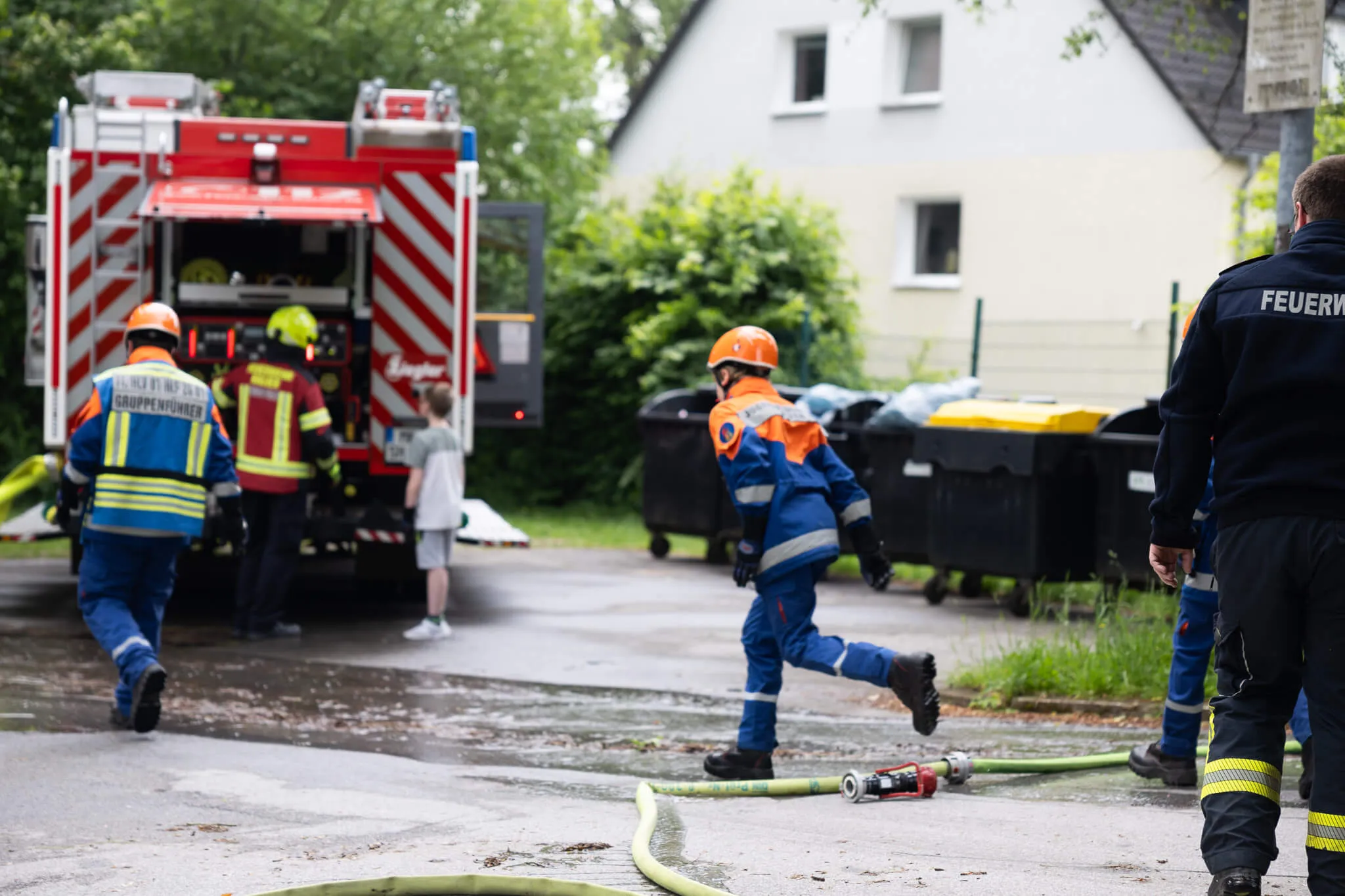 Feuerwehrleute bei einer Übung oder einem Einsatz vor einem Wohnhaus, während im Hintergrund ein Schulfest stattfindet.