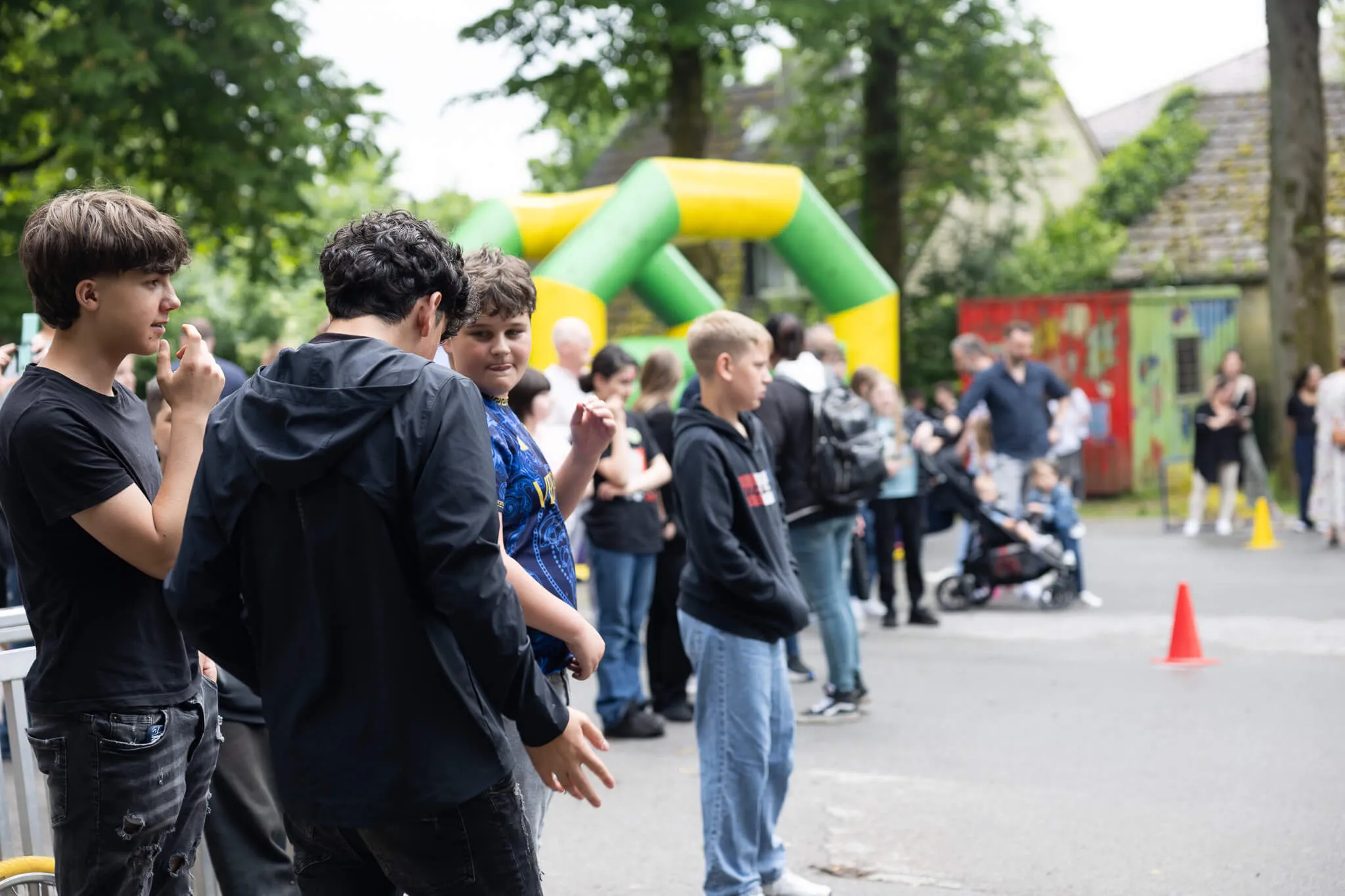 Mehrere Jugendliche stehen im Vordergrund, während im Hintergrund ein Schulfest mit einer Hüpfburg und anderen Besuchern stattfindet.