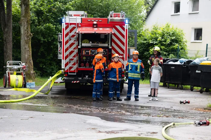 Feuerwehrleute in orangefarbenen Schutzanzügen stehen vor einem roten Feuerwehrauto, während Kinder zusehen.