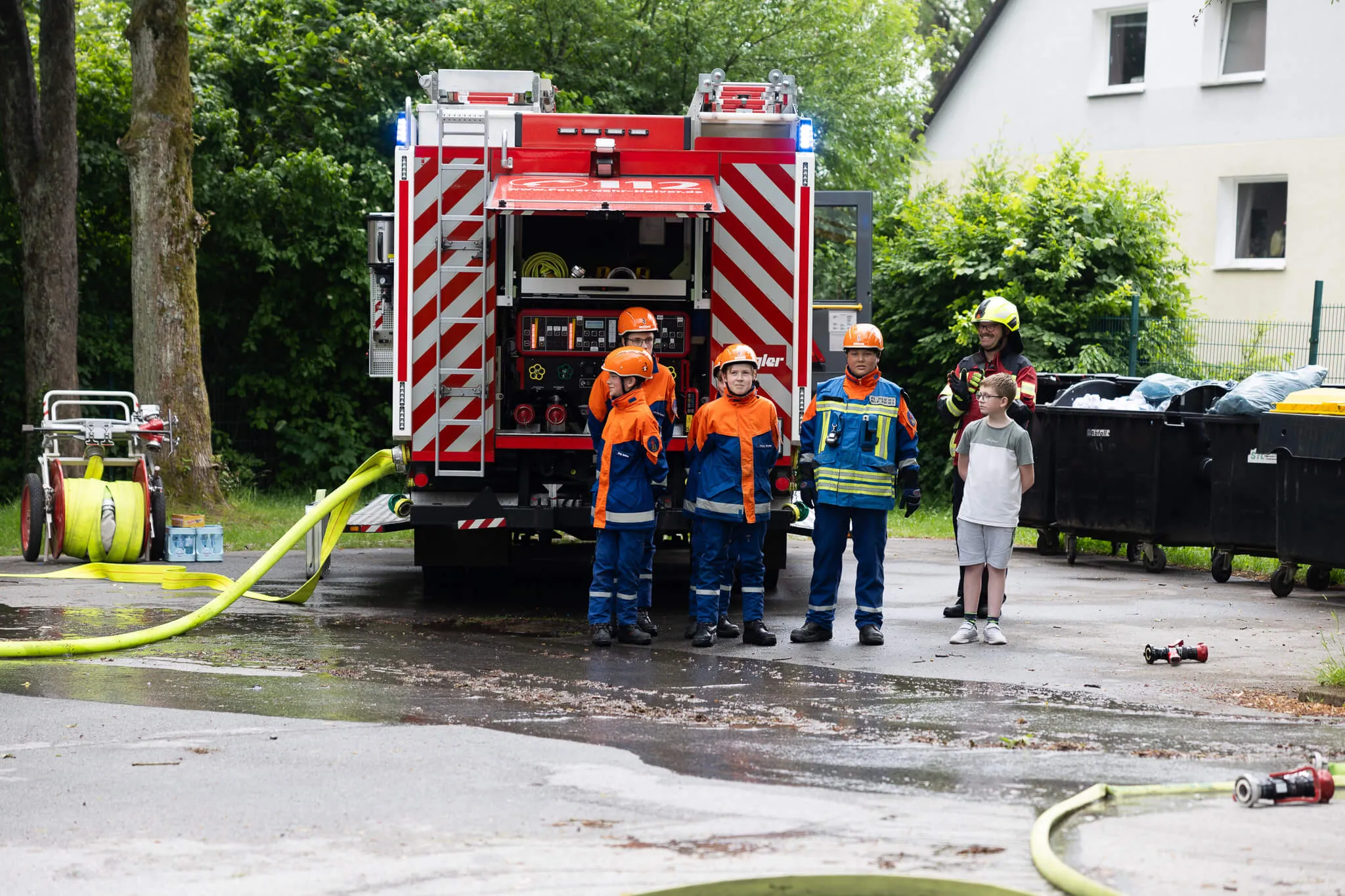 Feuerwehrleute in orangefarbenen Schutzanzügen stehen vor einem roten Feuerwehrauto, während Kinder zusehen.