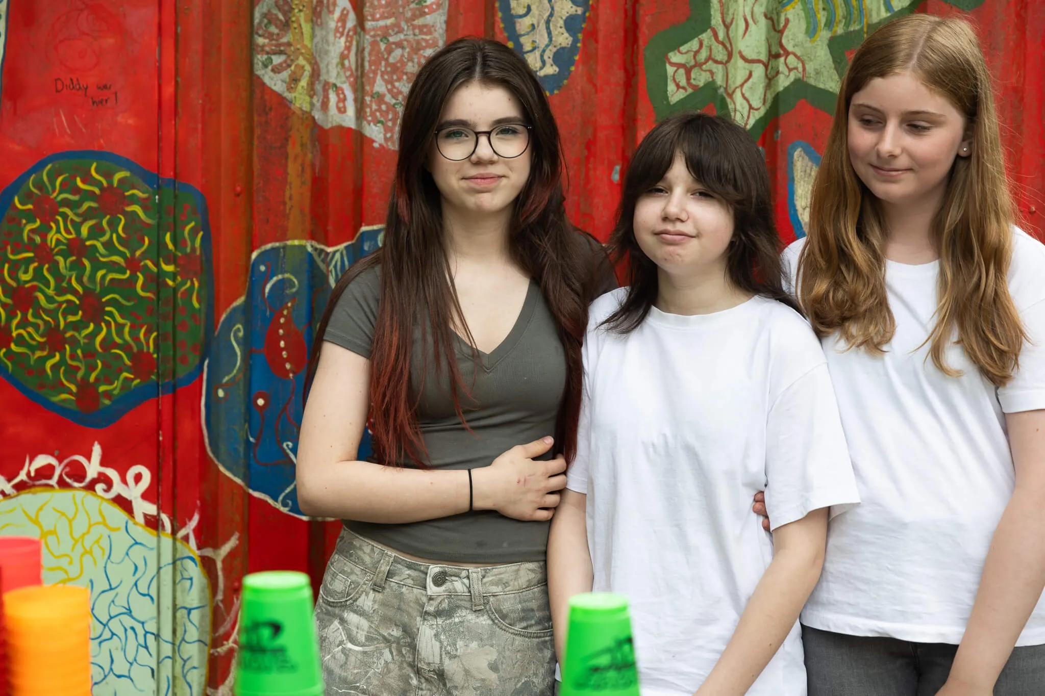 Drei junge Frauen stehen vor einer bunten Wand mit Graffiti und Plastikbechern auf einer Tischreihe. Sie lächeln in die Kamera.