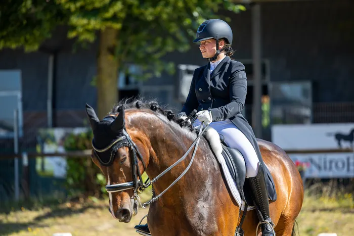 Eine Reiterin in schwarzer Jacke und Helm sitzt auf einem braunen Pferd. Das Pferd trägt eine schwarze Kopfhaube und ist mit Zügeln gezäumt. Im Hintergrund ist eine grüne Hecke und ein Gebäude zu sehen.