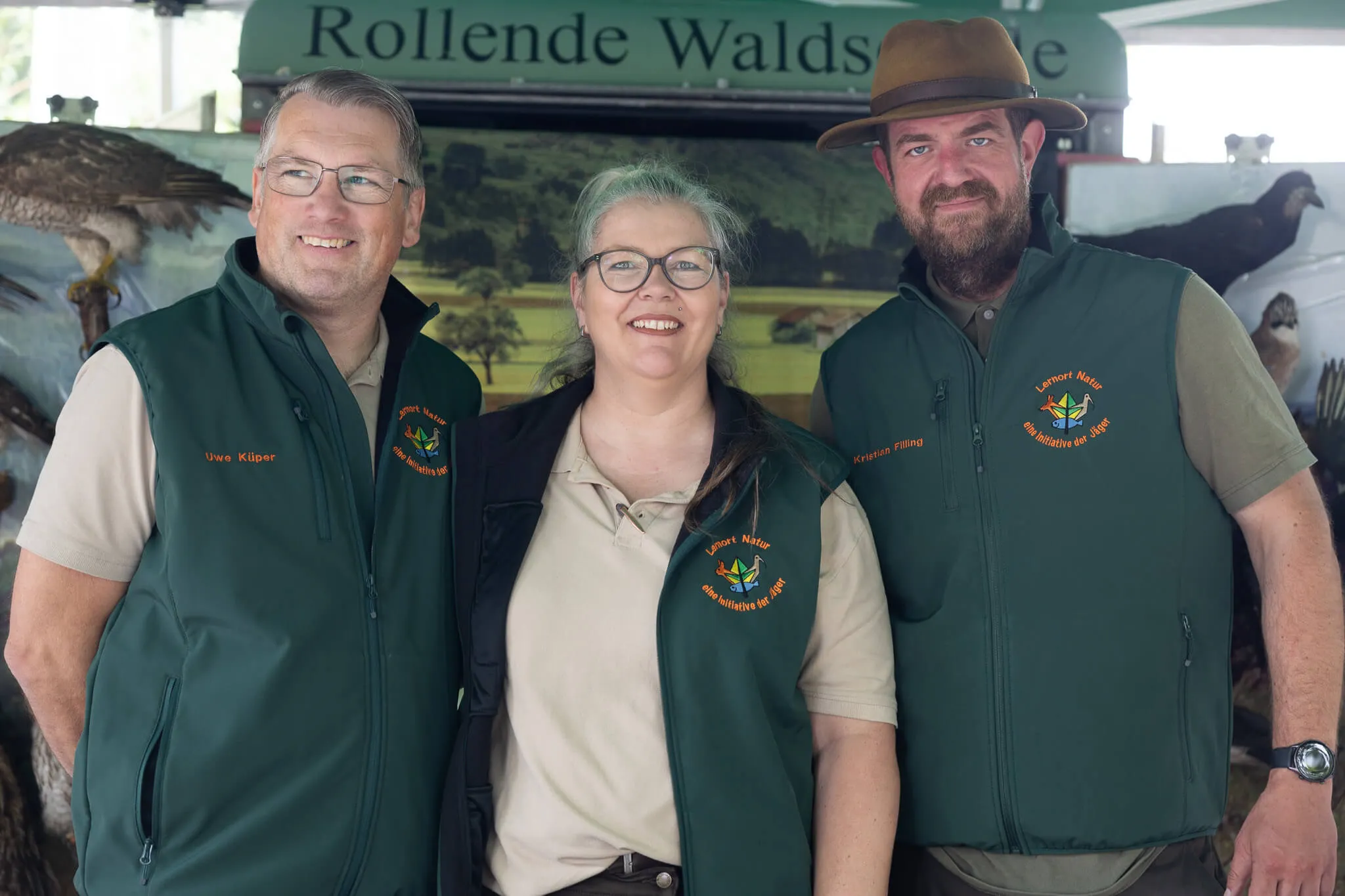 Drei Personen mit grünen Westen mit Logo „Lerner Natur“ stehen vor einem Banner mit der Aufschrift „Rollende Waldschule“.