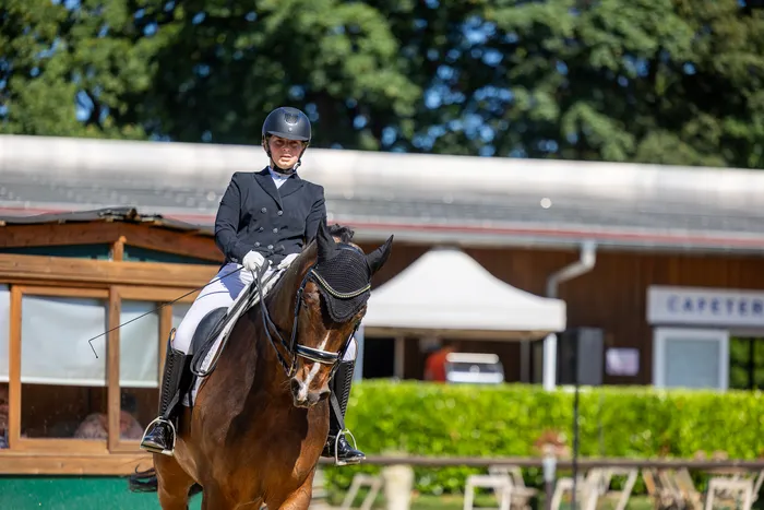 Eine Reiterin in schwarzer Turnierkleidung sitzt auf einem braunen Pferd und führt eine Dressurübung auf einem Reitplatz vor. Im Hintergrund sind Zelte und Gebäude zu sehen.