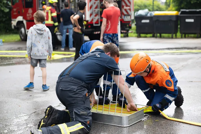 Ein Feuerwehrmann erklärt einem Kind in voller Schutzkleidung die Funktionsweise eines Metallgitters über einem Gully. Im Hintergrund sind weitere Kinder und ein rotes Feuerwehrauto zu sehen.