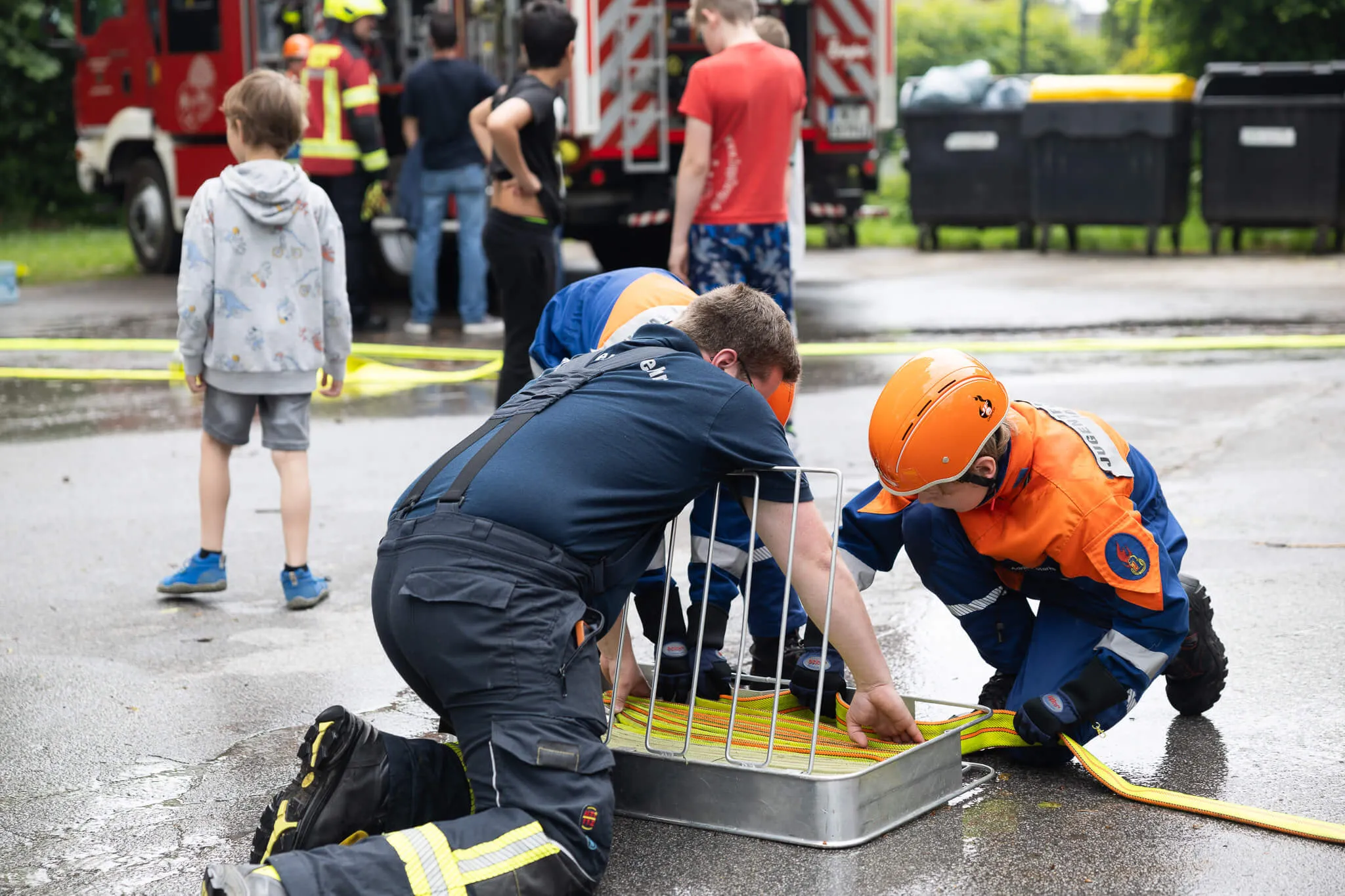 Ein Feuerwehrmann erklärt einem Kind in voller Schutzkleidung die Funktionsweise eines Metallgitters über einem Gully. Im Hintergrund sind weitere Kinder und ein rotes Feuerwehrauto zu sehen.