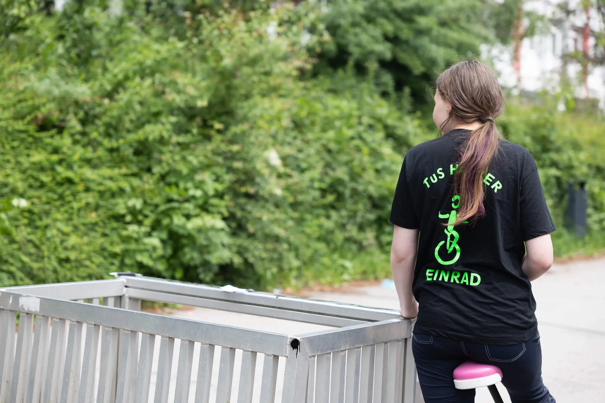 Frau mit langen, braunen Haaren steht mit dem Rücken zum Betrachter an einer Metallbarriere, trägt ein schwarzes T-Shirt mit dem Aufdruck 'TUS Hamer Einrad'.