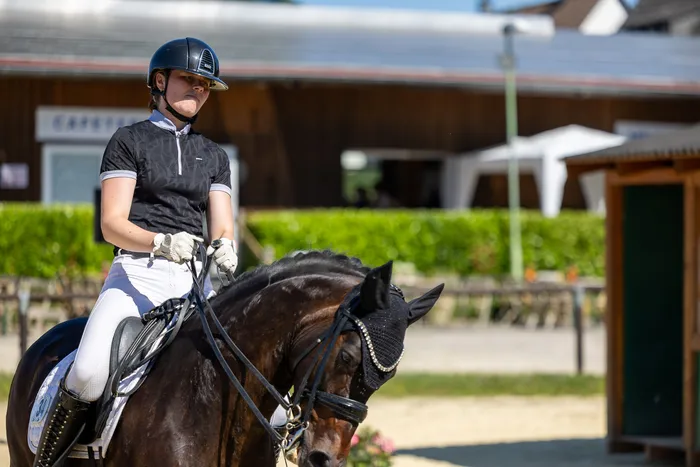 Eine Reiterin in schwarzer Kleidung und weißer Hose sitzt auf einem schwarzen Pferd. Das Pferd trägt eine Fliegenhaube und die Reiterin einen Helm. Im Hintergrund sind Gebäude und grüne Bäume zu sehen.
