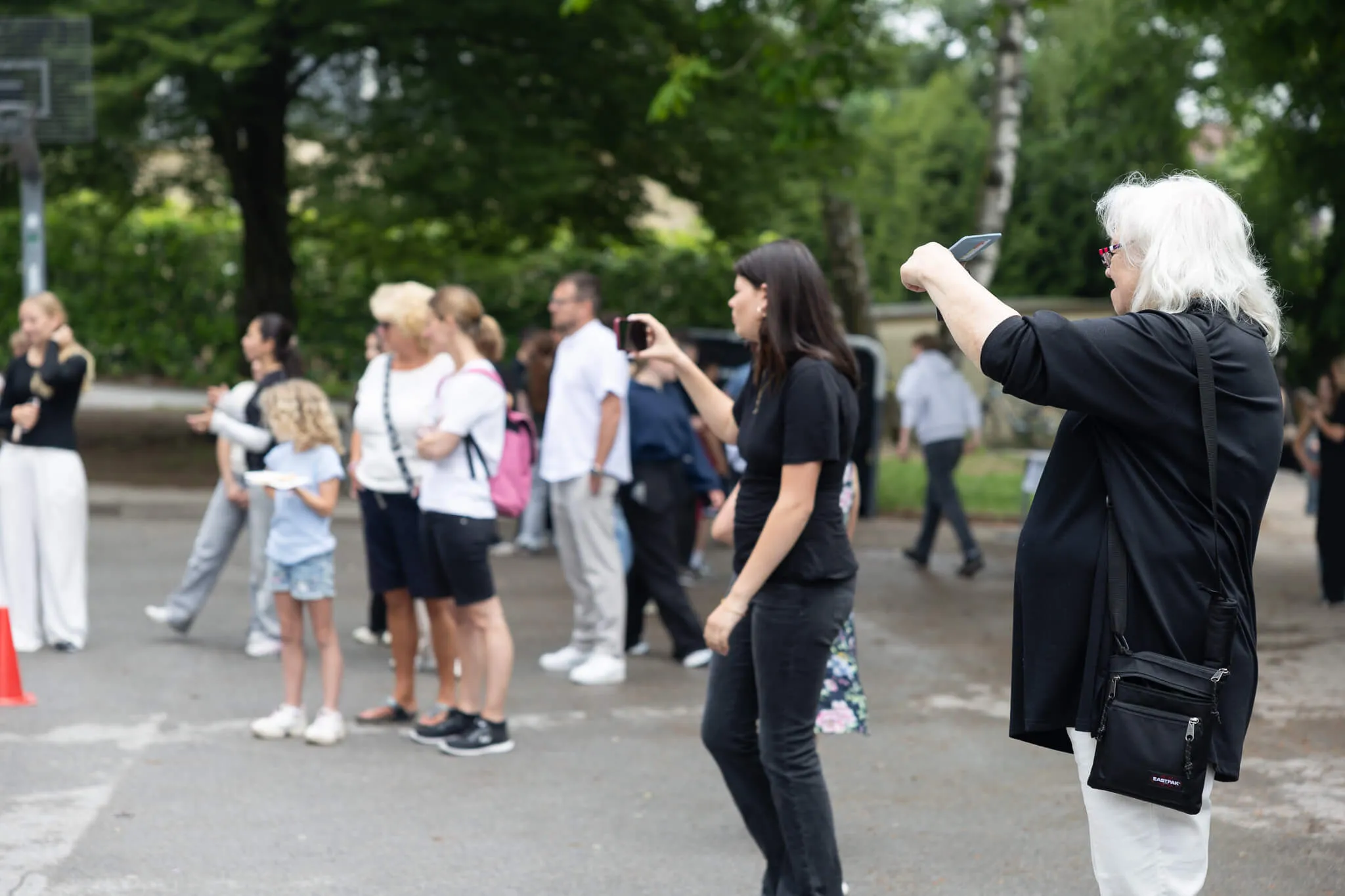 Eine Frau mit weißen Haaren fotografiert mit ihrem Smartphone eine Gruppe von Menschen, darunter Kinder, auf einem Schulfest.