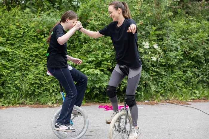 Zwei junge Frauen üben gemeinsam das Einradfahren auf einem Parkplatz, eine stabilisiert die andere.