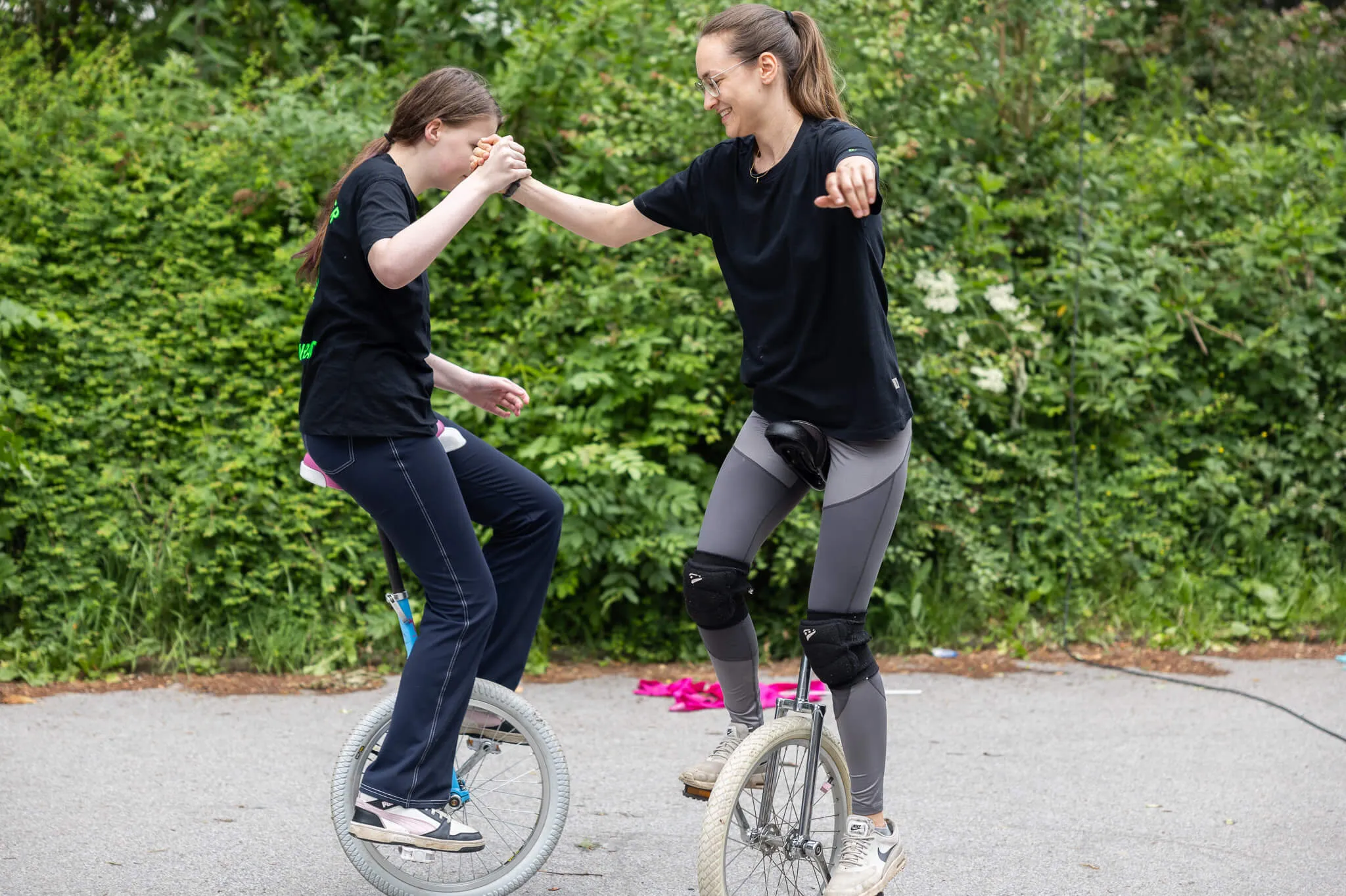 Zwei junge Frauen üben gemeinsam das Einradfahren auf einem Parkplatz, eine stabilisiert die andere.