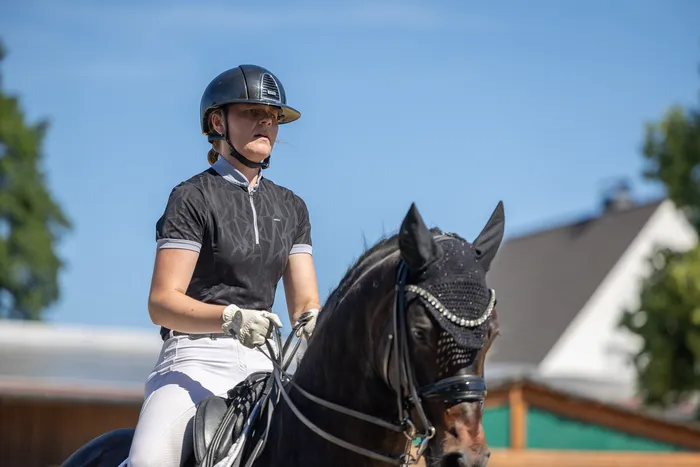 Eine Reiterin in weißer Hose und schwarzem Poloshirt sitzt auf einem schwarzen Pferd mit Zierde am Kopf. Sie trägt einen Helm und Handschuhe und blickt nach rechts. Im Hintergrund sind Bäume und ein Haus zu sehen.