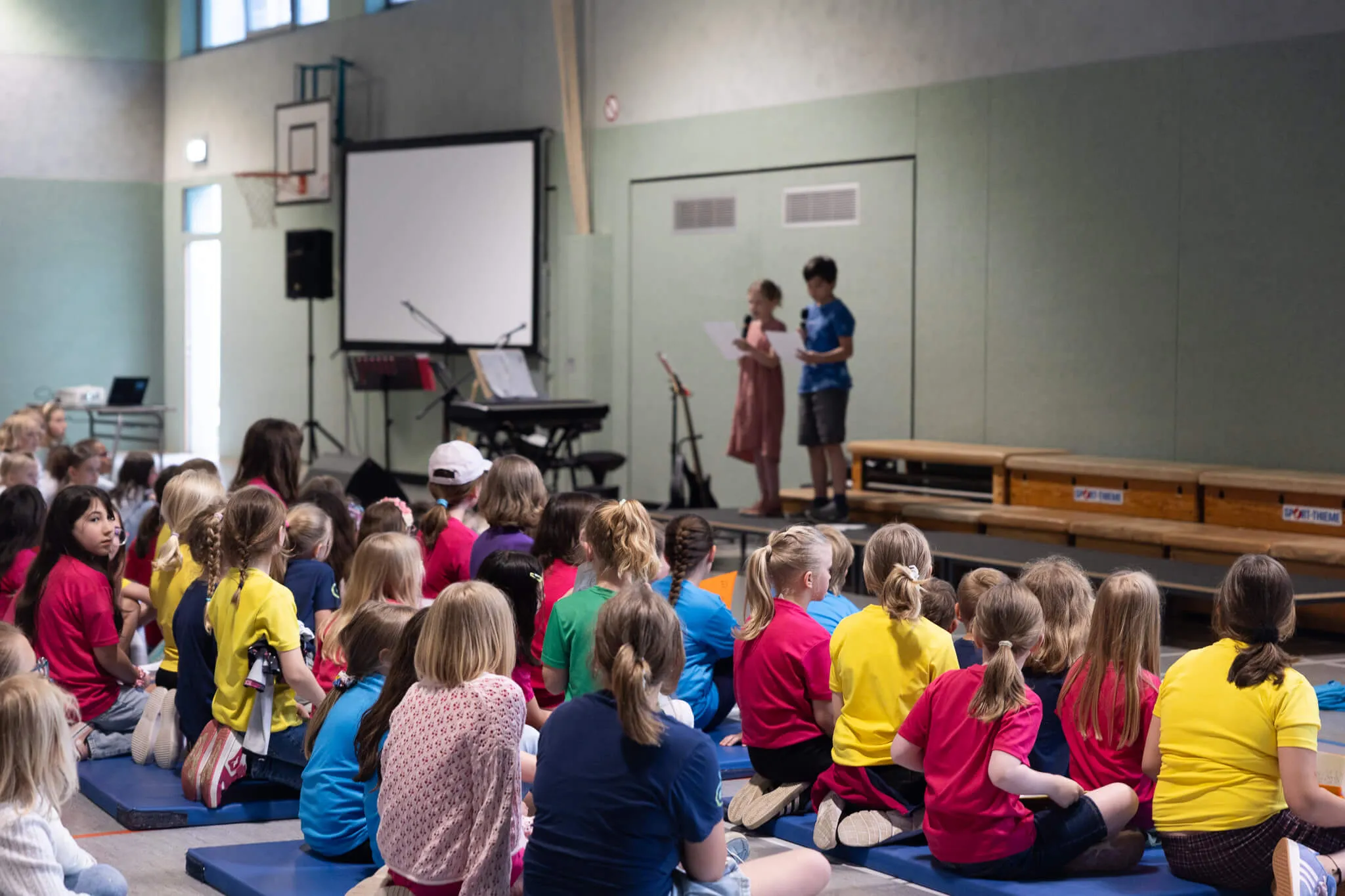 Schüler sitzen auf Matten in einer Turnhalle und hören einem Mitschüler zu, der auf einer kleinen Bühne etwas vorliest.