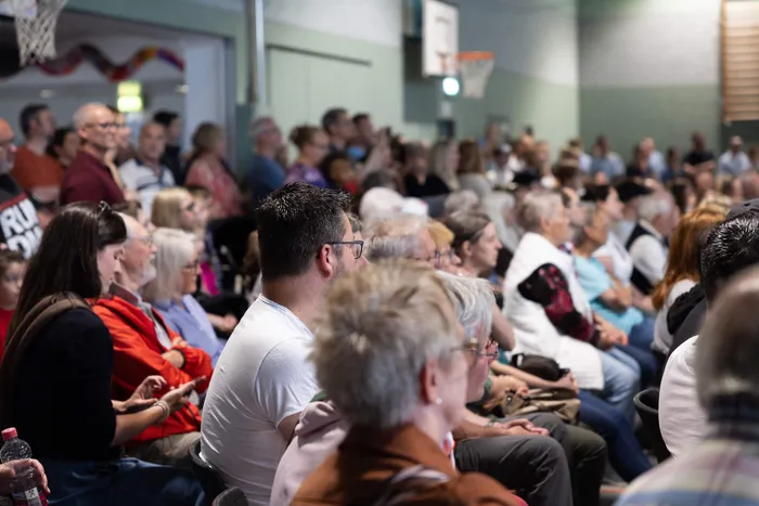 Zuschauer sitzen auf Stühlen in einer Turnhalle und beobachten eine Veranstaltung. Im Hintergrund sind Basketballkörbe und Fenster zu sehen.