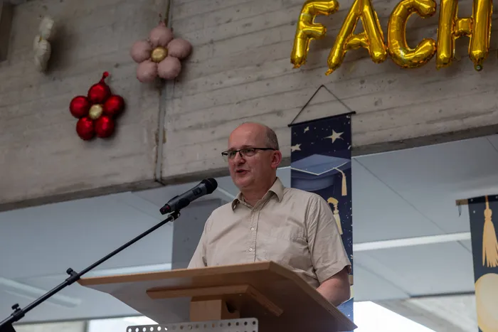 Ein Mann mit Glatze und Brille spricht in ein Mikrofon an einem Rednerpult. Im Hintergrund hängen goldene Buchstaben, die „FACH“ ergeben, sowie ein Banner mit einem Diplomhut und Blumen.