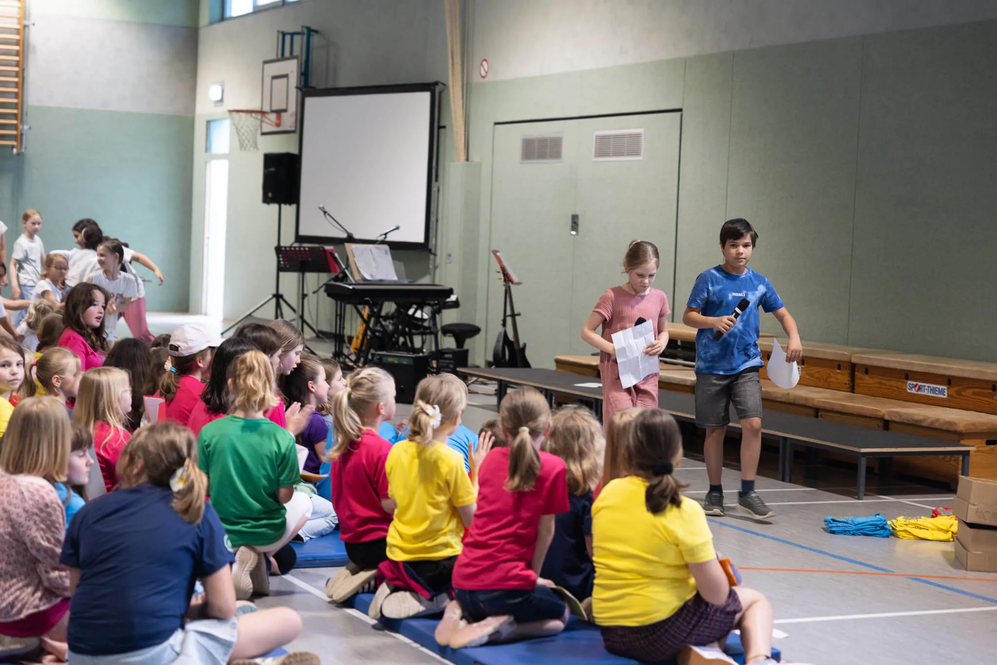 Ein Junge steht vor einer Gruppe von Kindern in einer Turnhalle und hält Papiere in der Hand. Im Hintergrund eine Bühne und eine Leinwand.