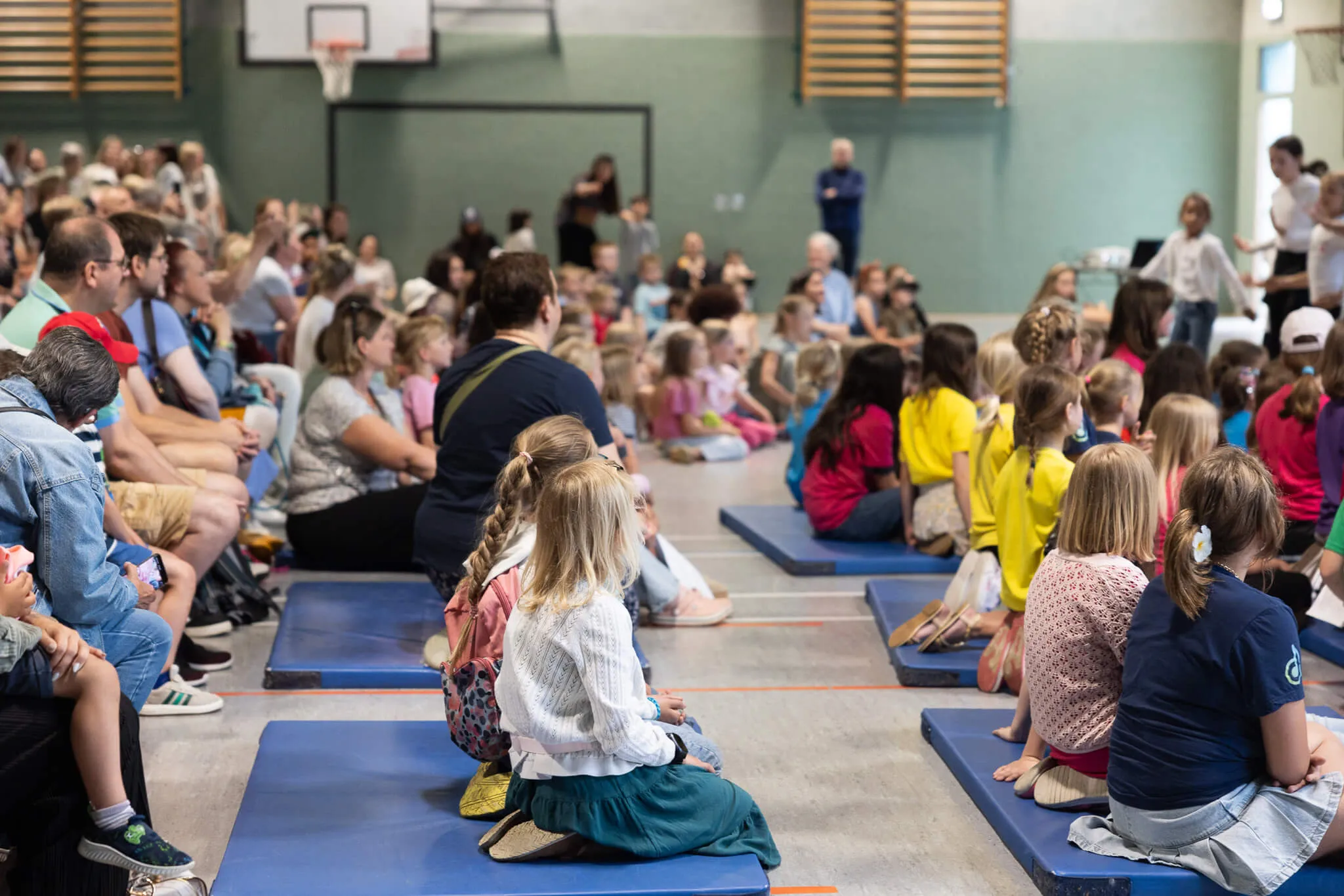 Große Gruppe von Kindern und Eltern sitzt auf blauen Matten in einer Turnhalle und blickt nach vorne, wo ein Mann am Rednerpult steht.