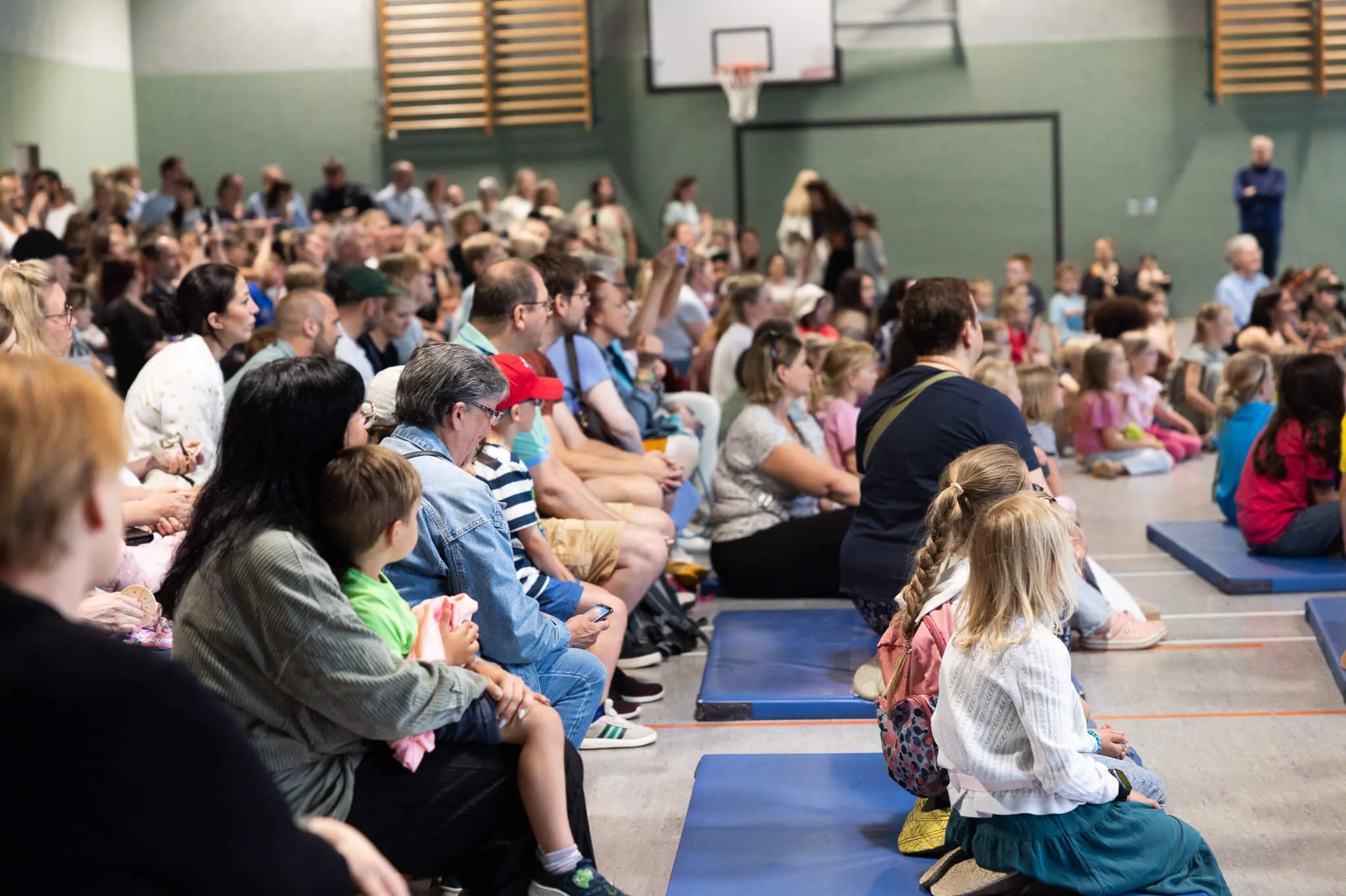 Große Menschenmenge sitzt auf blauen Matten in einer Turnhalle und blickt nach vorne, im Hintergrund ein Basketballkorb.