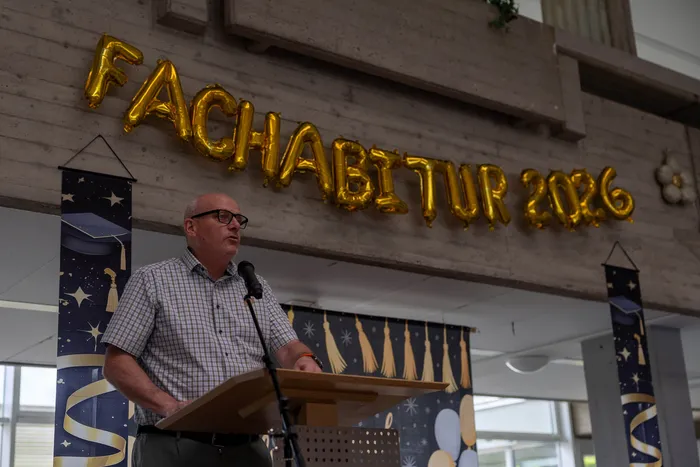 Ein Mann mit Brille spricht an einem Rednerpult in eine Mikrofon. Im Hintergrund hängt ein Banner mit der Aufschrift „Fachabitur 2026“.