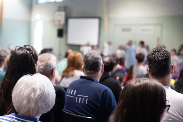 Zuschauer sitzen auf Stühlen in einer Turnhalle und blicken auf eine Gruppe von Menschen, die im Vordergrund stehen. Im Hintergrund eine weiße Leinwand.