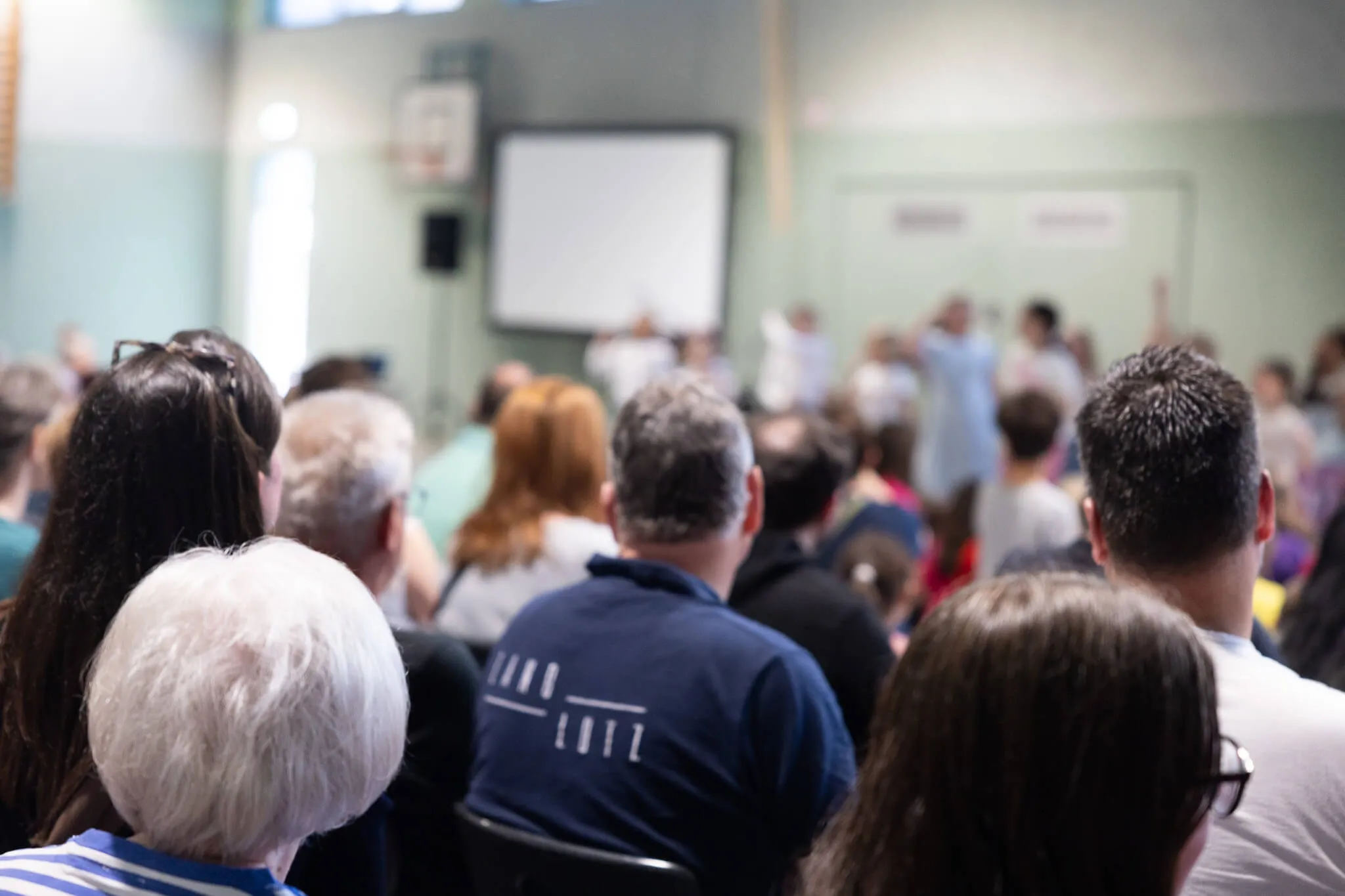Zuschauer sitzen auf Stühlen in einer Turnhalle und blicken auf eine Gruppe von Menschen, die im Vordergrund stehen. Im Hintergrund eine weiße Leinwand.