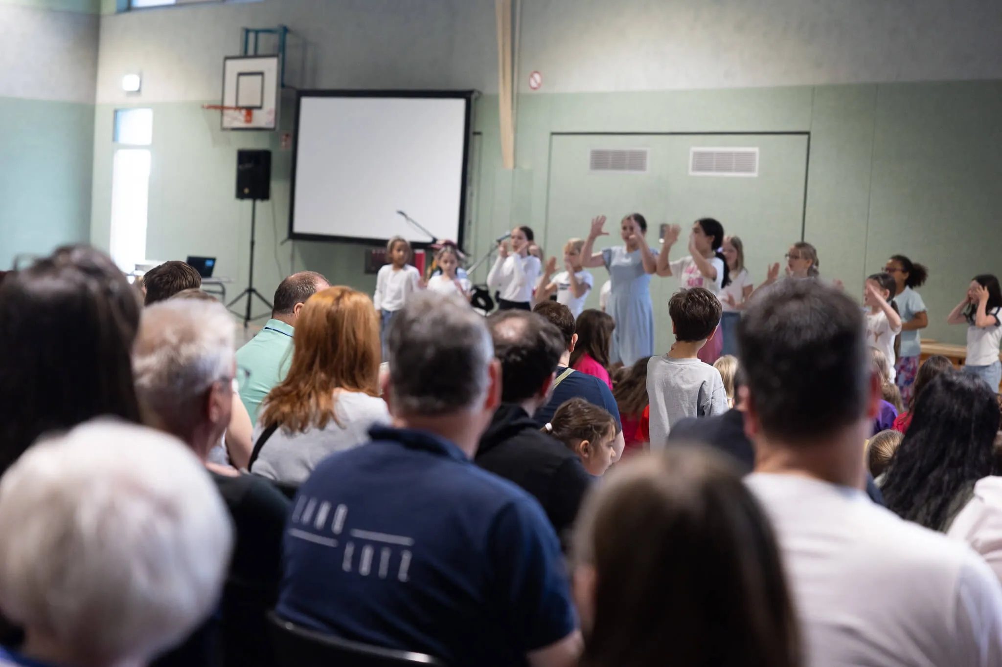 Eine Gruppe von Kindern führt vor einem Publikum in einer Turnhalle einen Tanz auf. Im Hintergrund eine leere Leinwand und ein Basketballkorb.