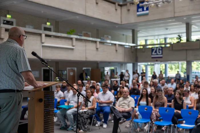 Ein Mann spricht an einem Pult vor einer großen Gruppe von Menschen, die auf blauen Stühlen sitzen. Im Hintergrund ist ein helles, modernes Gebäude mit Glasfenstern zu sehen.