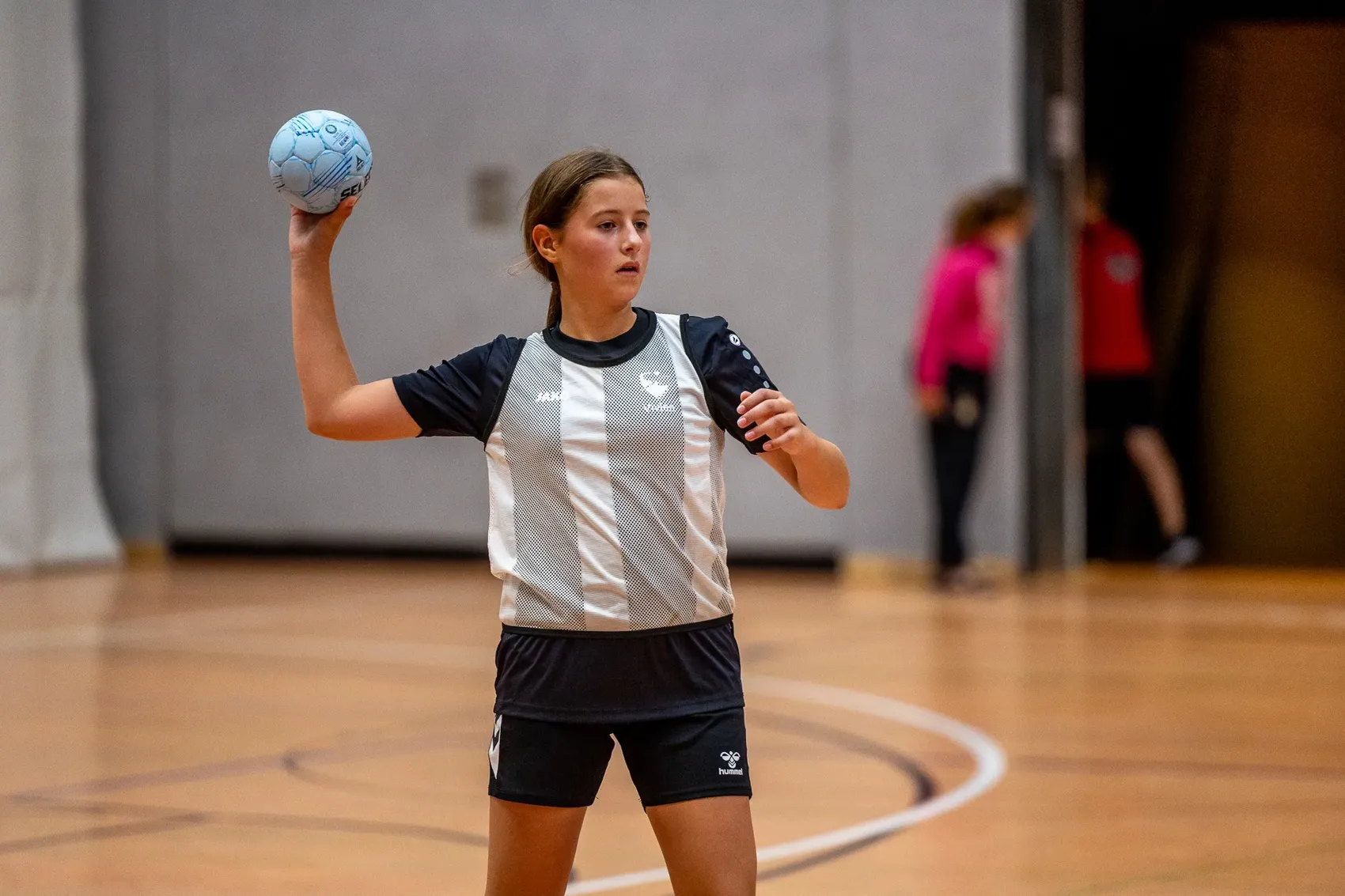 Ein Mädchen in einem schwarz-weiß gestreiften Trikot hält einen blauen Handball in der erhobenen Hand bereit zum Wurf.