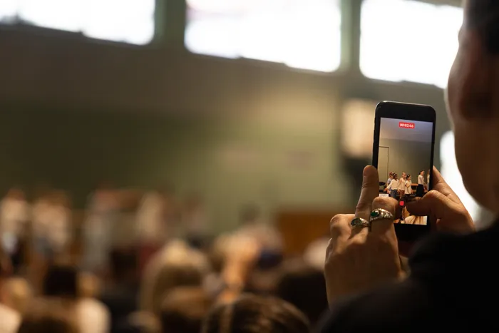 Eine Person filmt mit einem Smartphone eine Bühne, auf der drei Musiker spielen. Im Hintergrund sind verschwommene Zuschauer in einer Halle zu sehen.