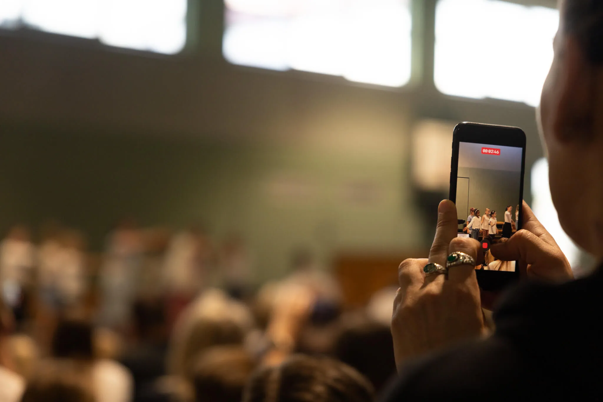 Eine Person filmt mit einem Smartphone eine Bühne, auf der drei Musiker spielen. Im Hintergrund sind verschwommene Zuschauer in einer Halle zu sehen.