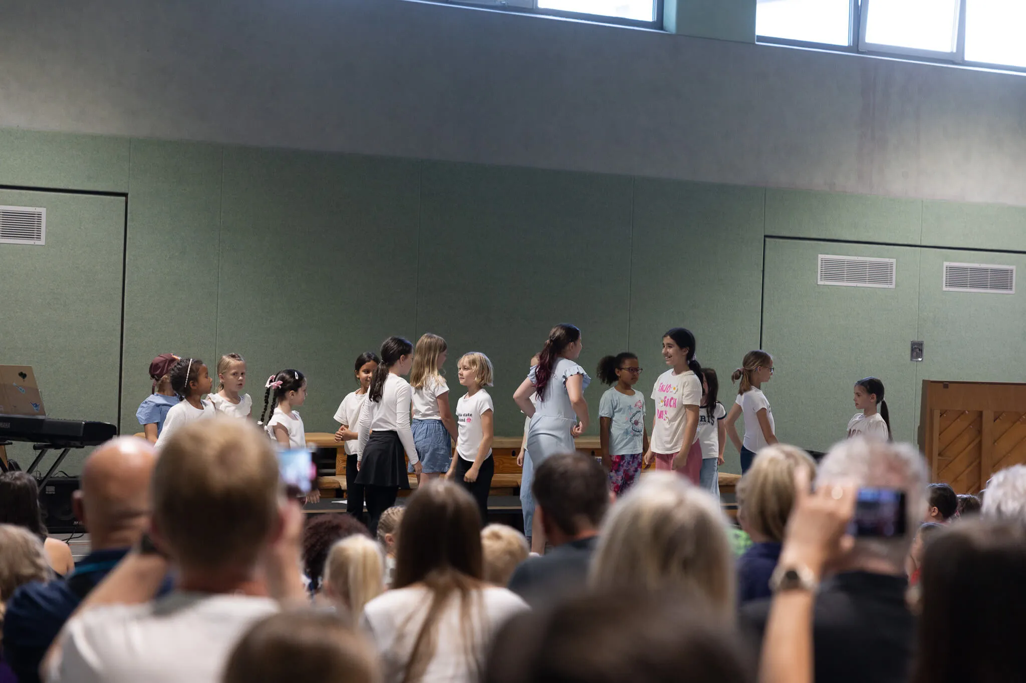 Eine Gruppe von Kindern steht auf einer Bühne und lächelt in die Menge. Im Hintergrund sitzen und stehen zahlreiche Zuschauer in einem hellen Raum.