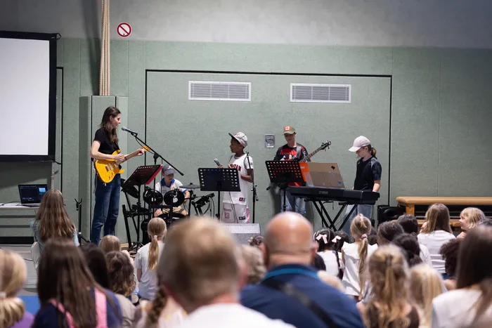 Eine Band aus Schüler*innen spielt vor einem Publikum in einer Turnhalle. Im Vordergrund eine Person mit grauem Haar, die eine E-Gitarre spielt, daneben ein Kind mit Basecap am Schlagzeug und ein weiteres Kind mit Keyboard.