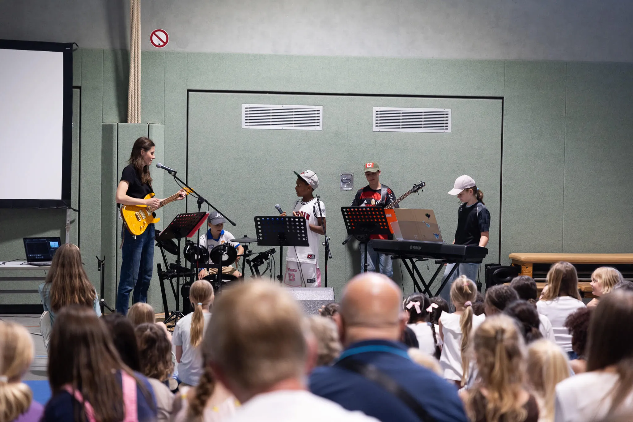 Eine Band aus Schüler*innen spielt vor einem Publikum in einer Turnhalle. Im Vordergrund eine Person mit grauem Haar, die eine E-Gitarre spielt, daneben ein Kind mit Basecap am Schlagzeug und ein weiteres Kind mit Keyboard.