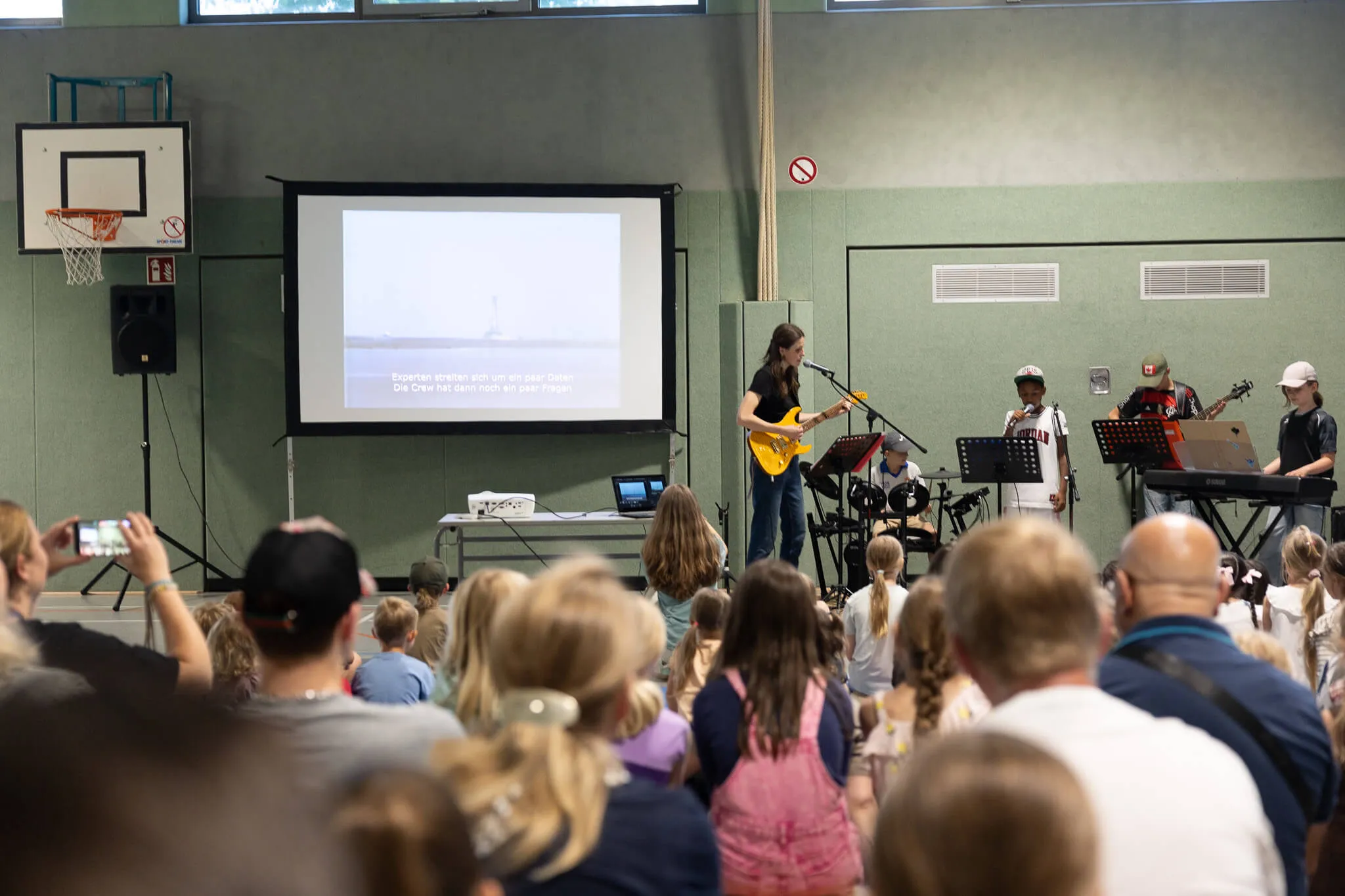 Frau spielt Gitarre vor Publikum in einer Turnhalle, während auf einer Leinwand ein Bild von einem Segelboot zu sehen ist. Im Hintergrund weitere Musiker und ein Mann mit einer roten Kappe.