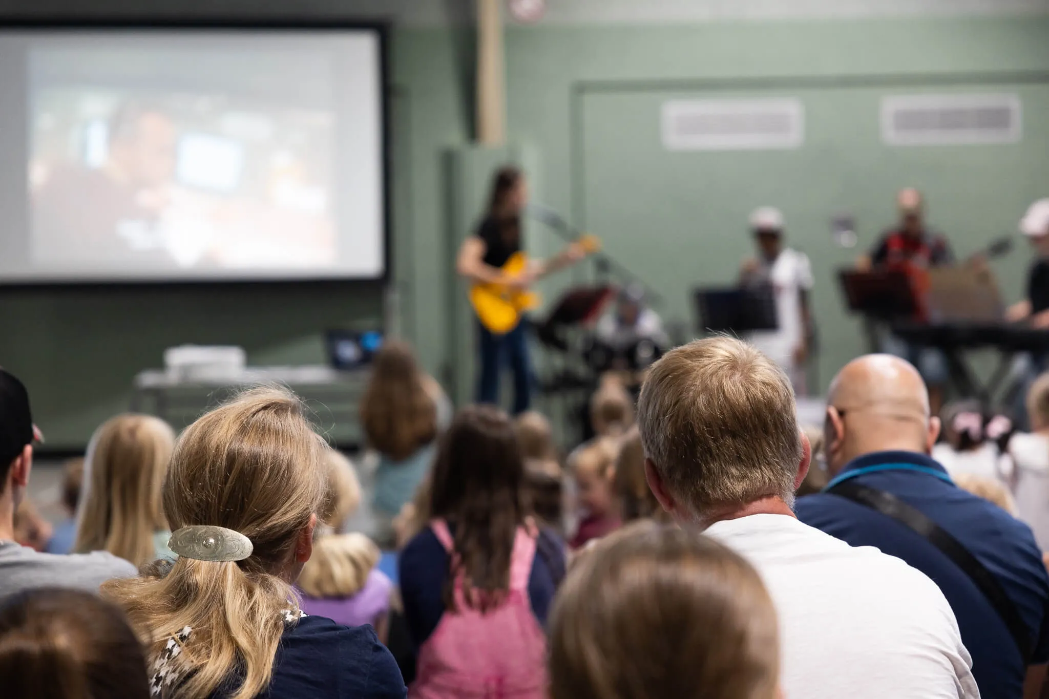 Publikum sitzt einer Musikerin mit Gitarre auf einer Bühne im neuen Musikschulgebäude der Lindenhofschule zugewandt. Auf einer Leinwand im Hintergrund ist ein Bild zu sehen.