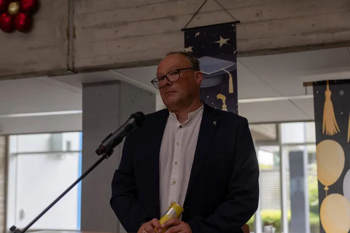 Ein Mann mit Brille und dunklem Anzug spricht in ein Mikrofon. Im Hintergrund sind Dekorationen mit dem Motiv einer Mütze zu sehen, die für einen Schulabschluss steht.